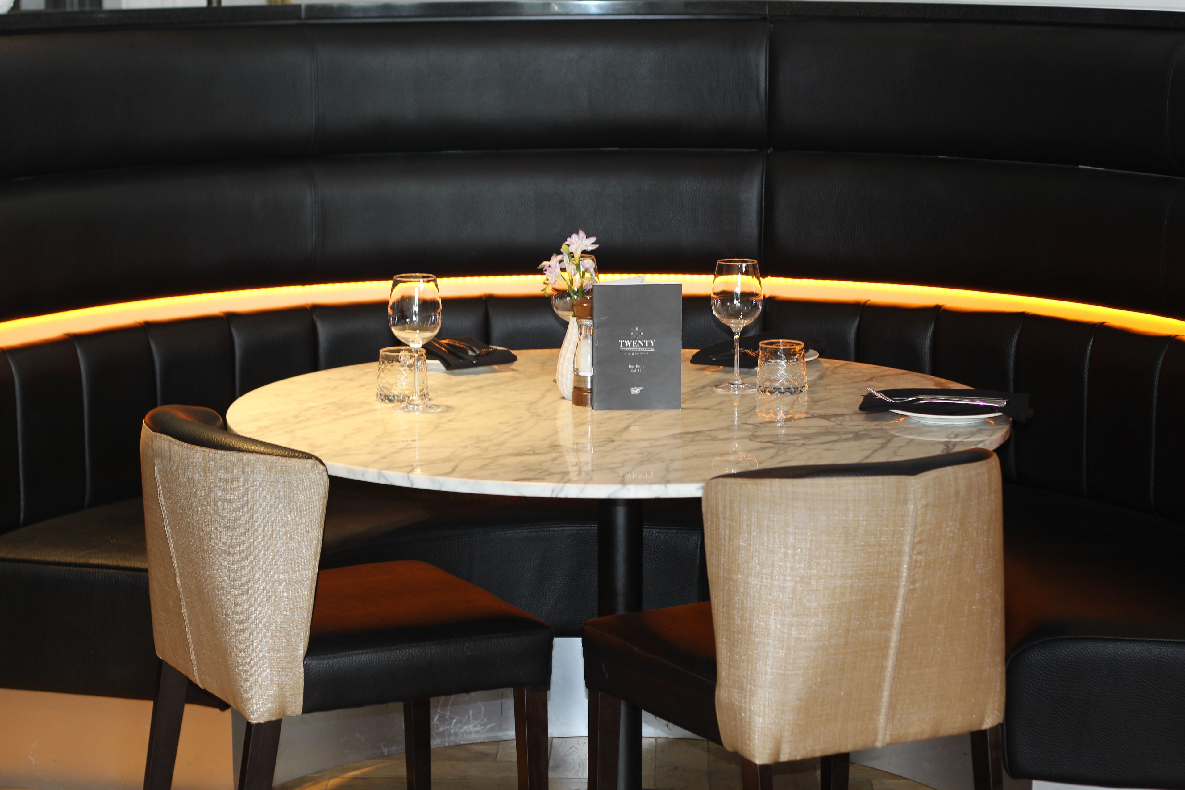 A round table with a marble top, surrounded by four upholstered chairs with a light fabric. A black menu stand with a menu titled 'TWENTY' is placed on the table.