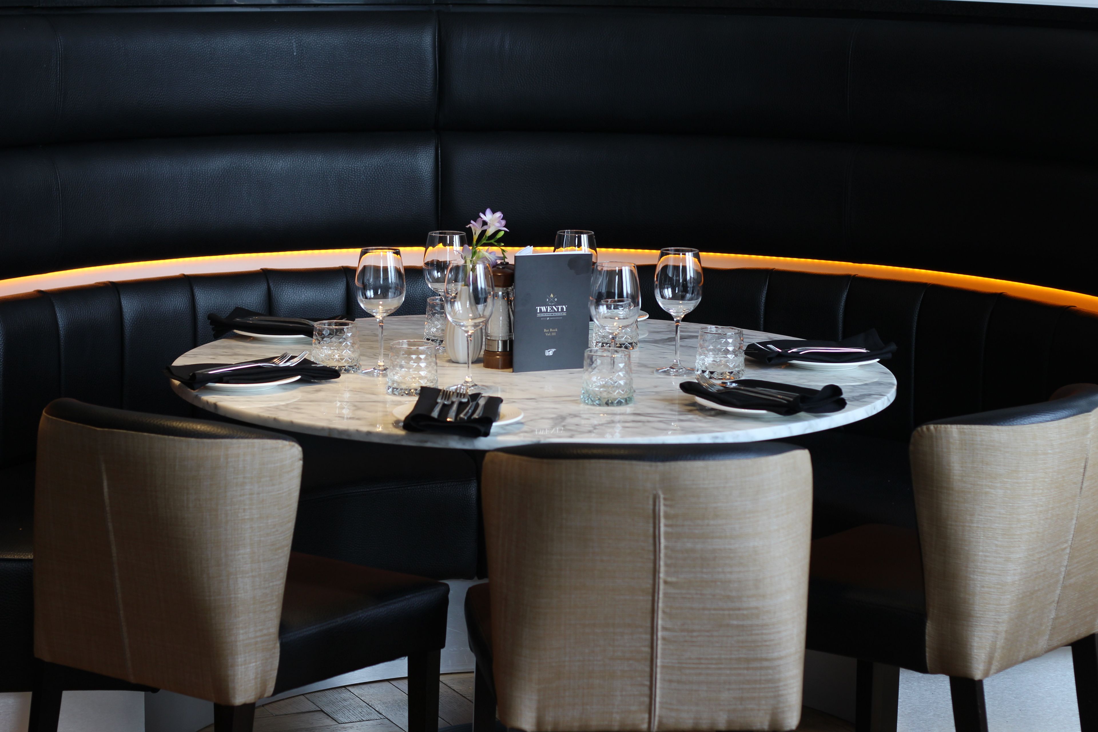 A round table with a marble top, surrounded by four upholstered chairs with a light fabric. A menu stand with a menu titled 'TWENTY' is placed in the center.