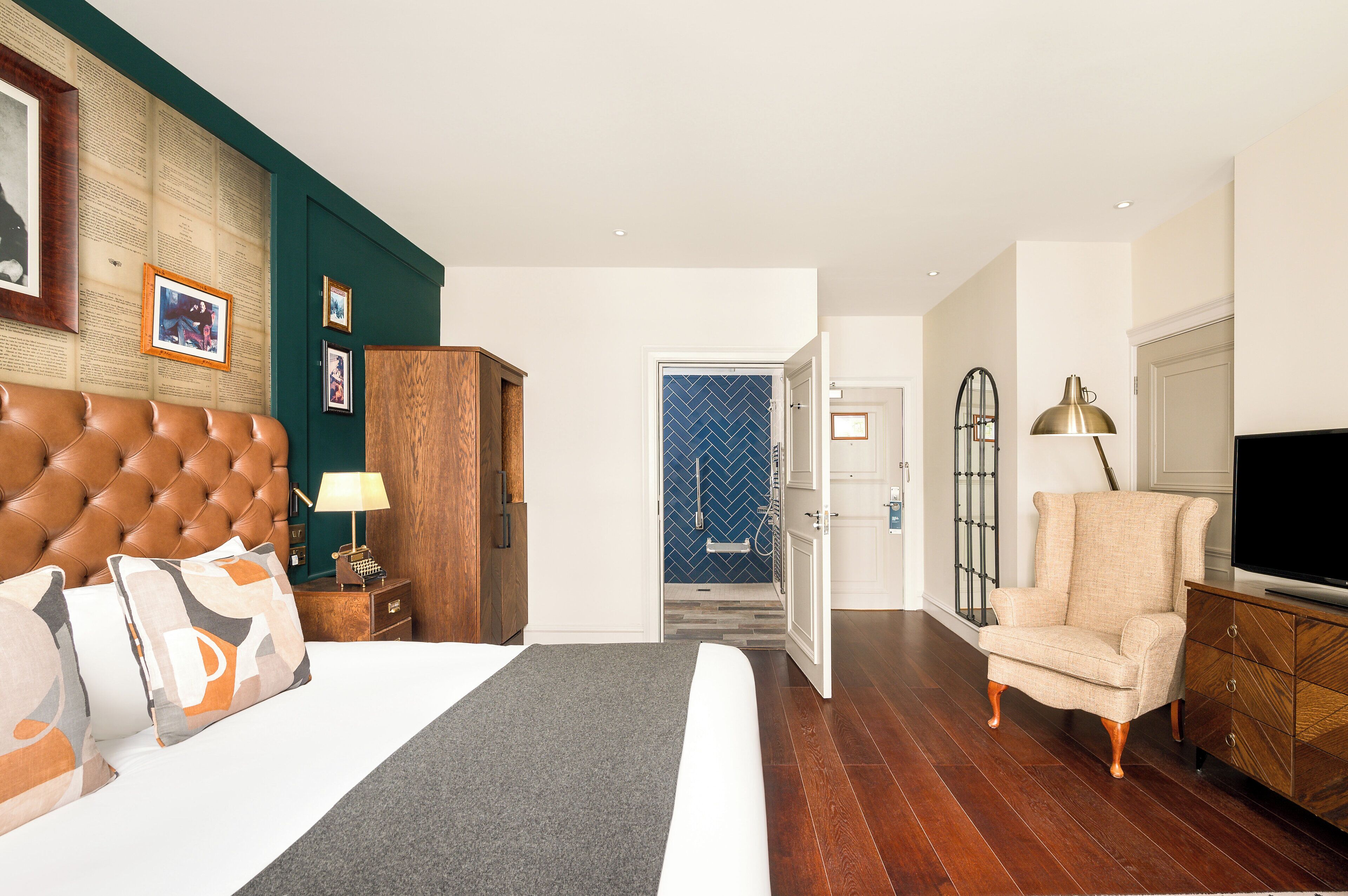 A hotel room with a large bed, a wooden headboard, a desk with a lamp, a television, and a seating area with a chair.