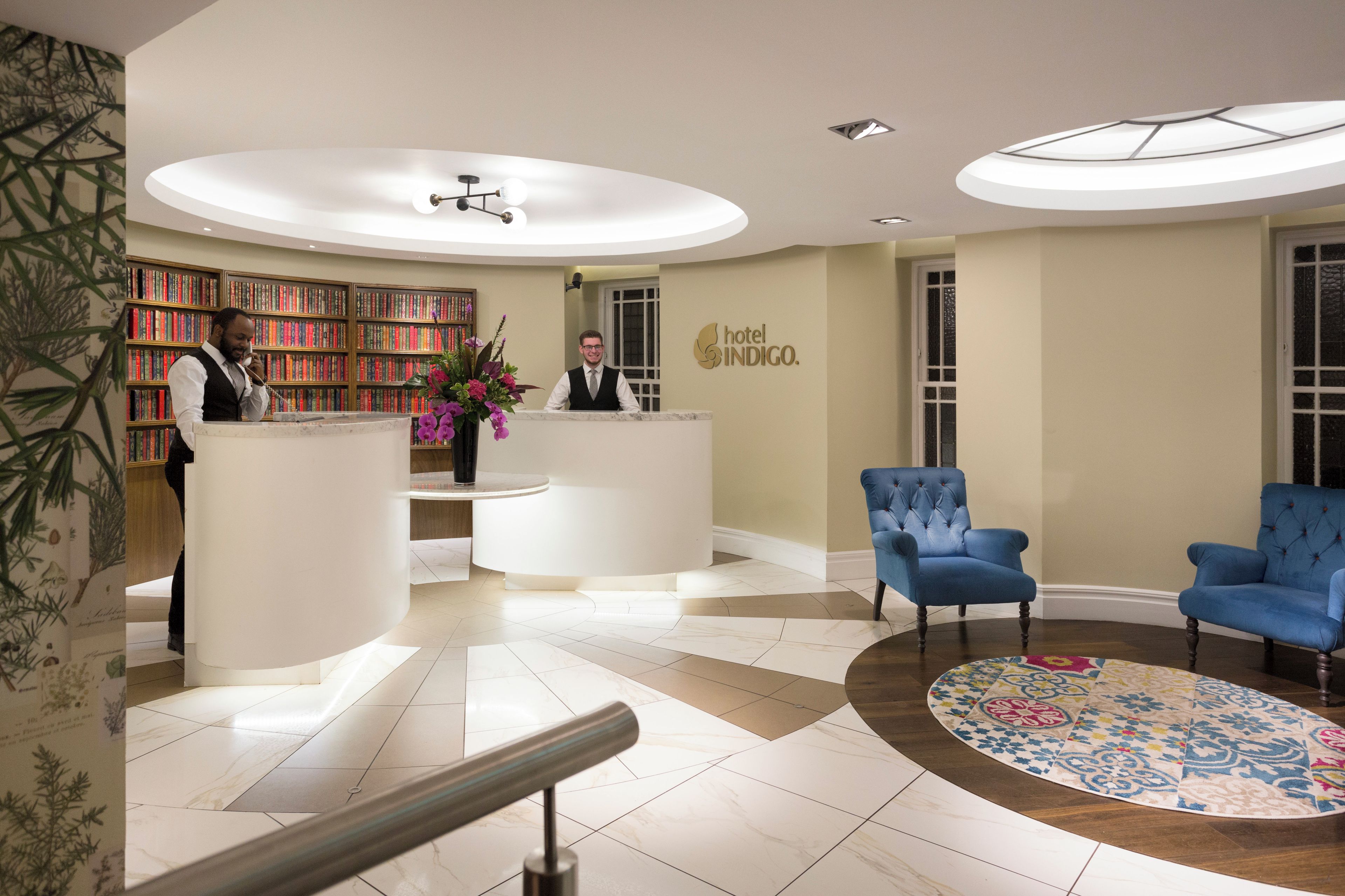 A hotel reception area with a reception desk, a bookshelf, a blue armchair, and a patterned rug.