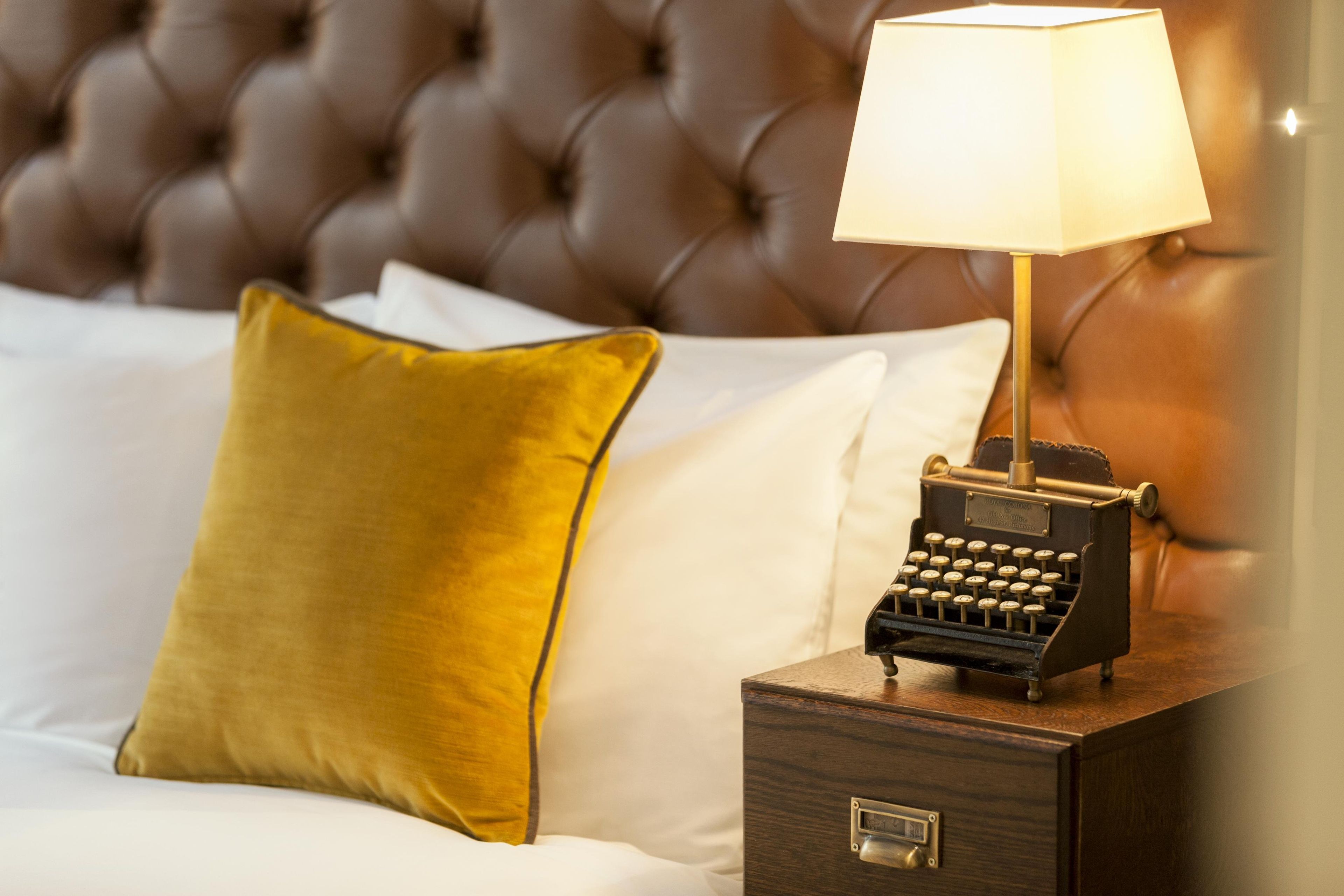 A bed with a tufted headboard, a yellow pillow, and a nightstand with a lamp and a typewriter.