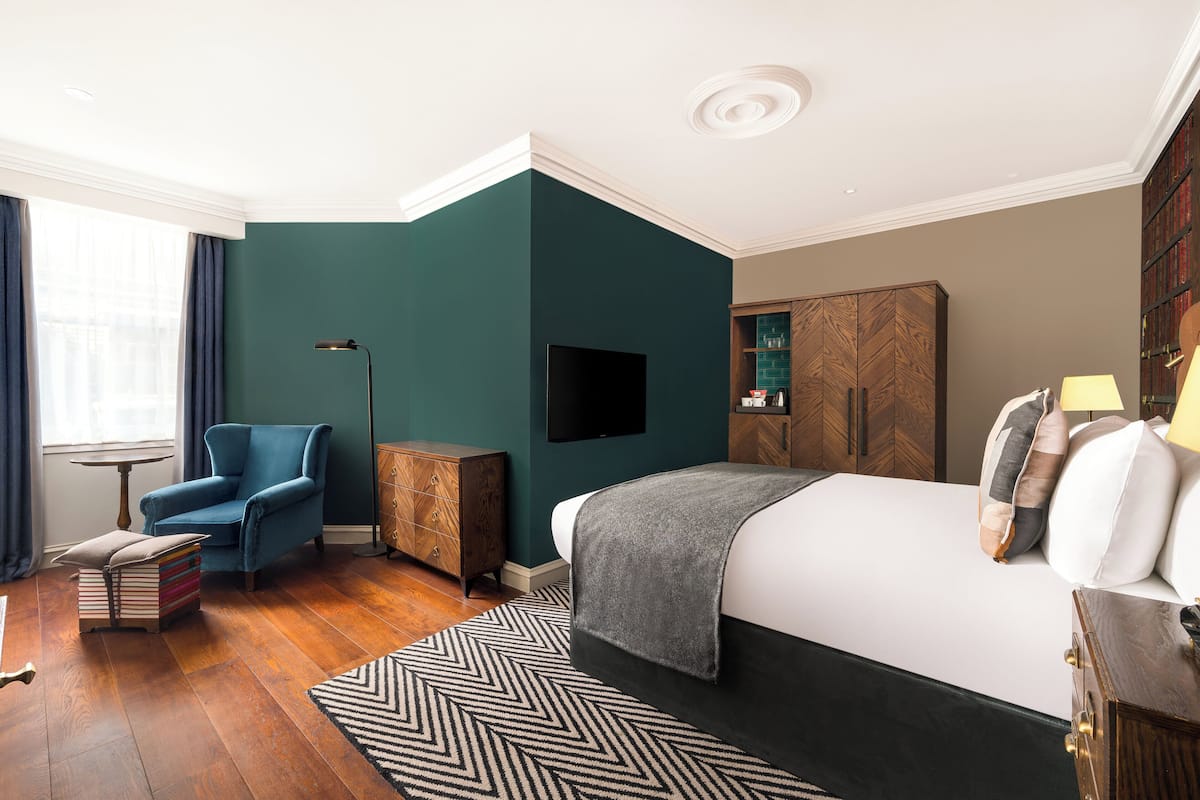 A hotel room with a bed, a blue armchair, a small table, a bookshelf, and a television.