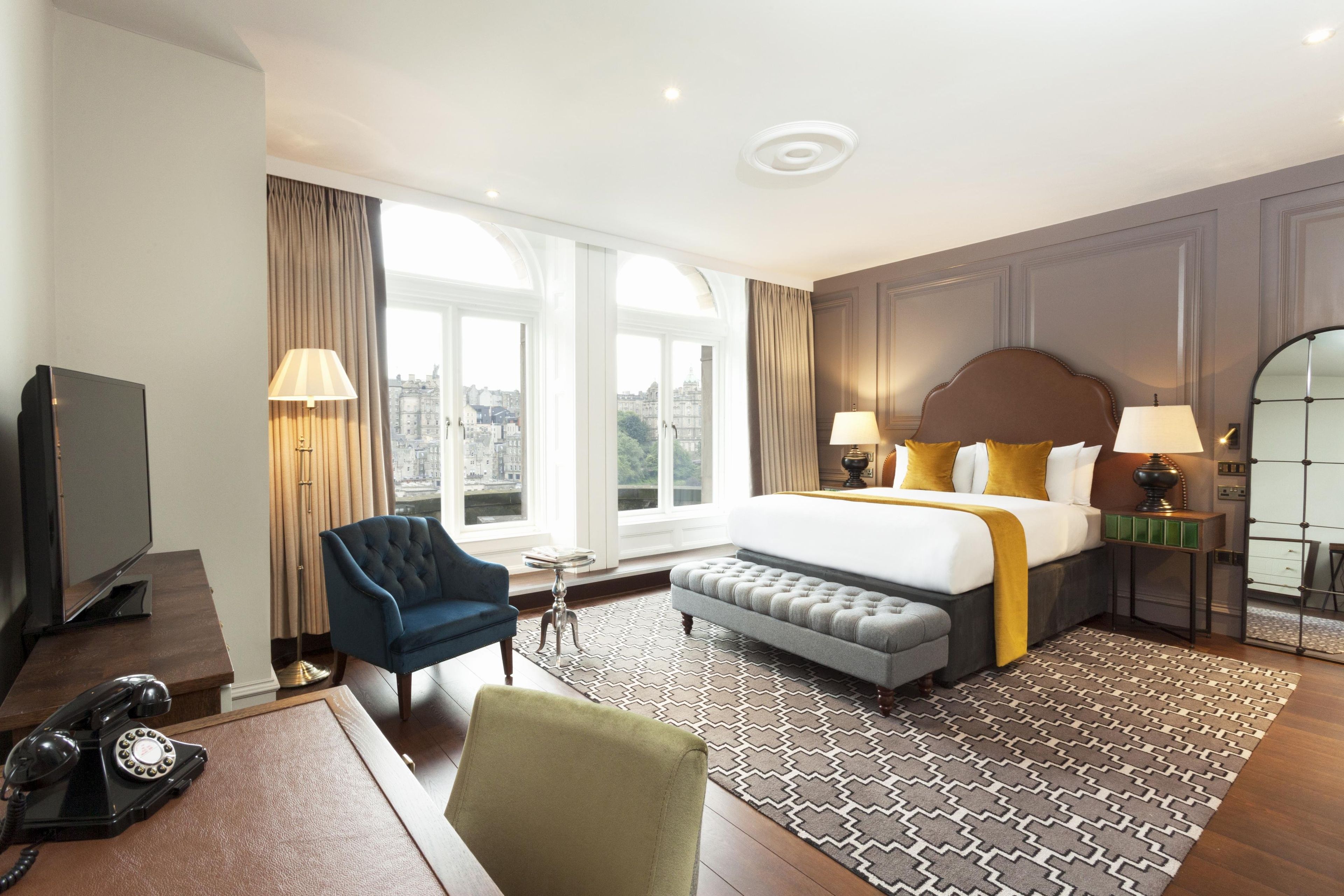 A hotel room with a bed, two lamps, a chair, a desk with a telephone, and a view of the city through large windows.