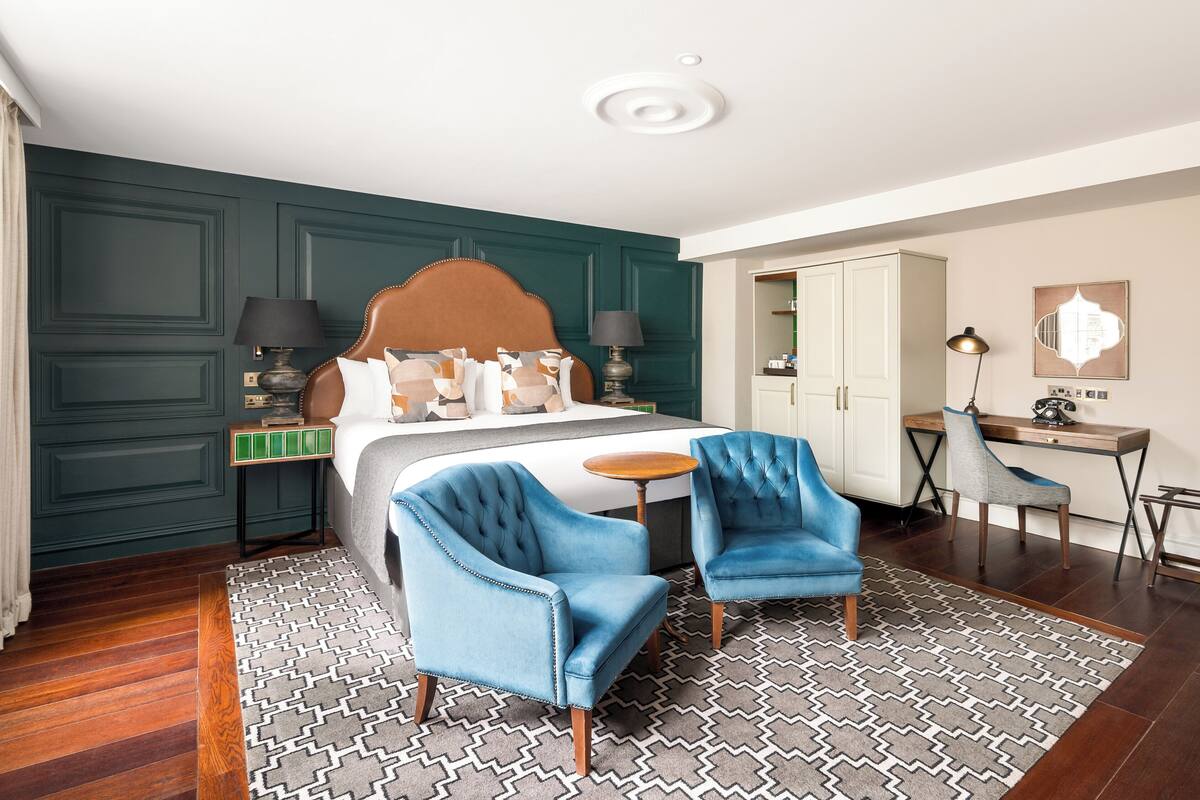A hotel room with a bed, two blue armchairs, a desk, and a wardrobe.