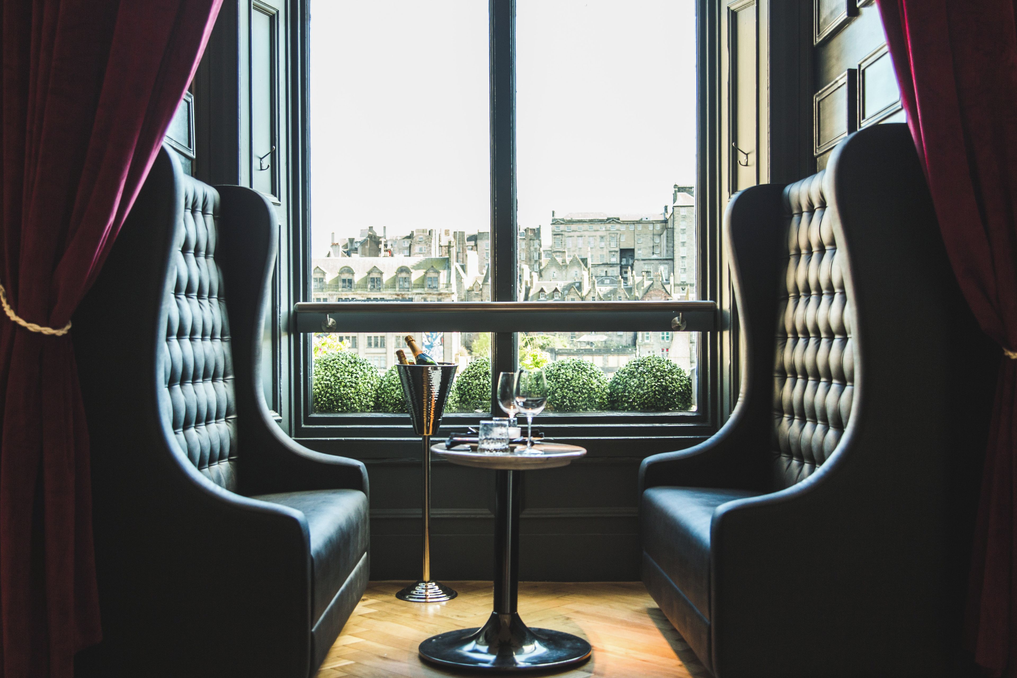 A corner seating area with two black upholstered chairs, a small round table with a bottle and a glass, and a view of buildings through a window with red curtains.