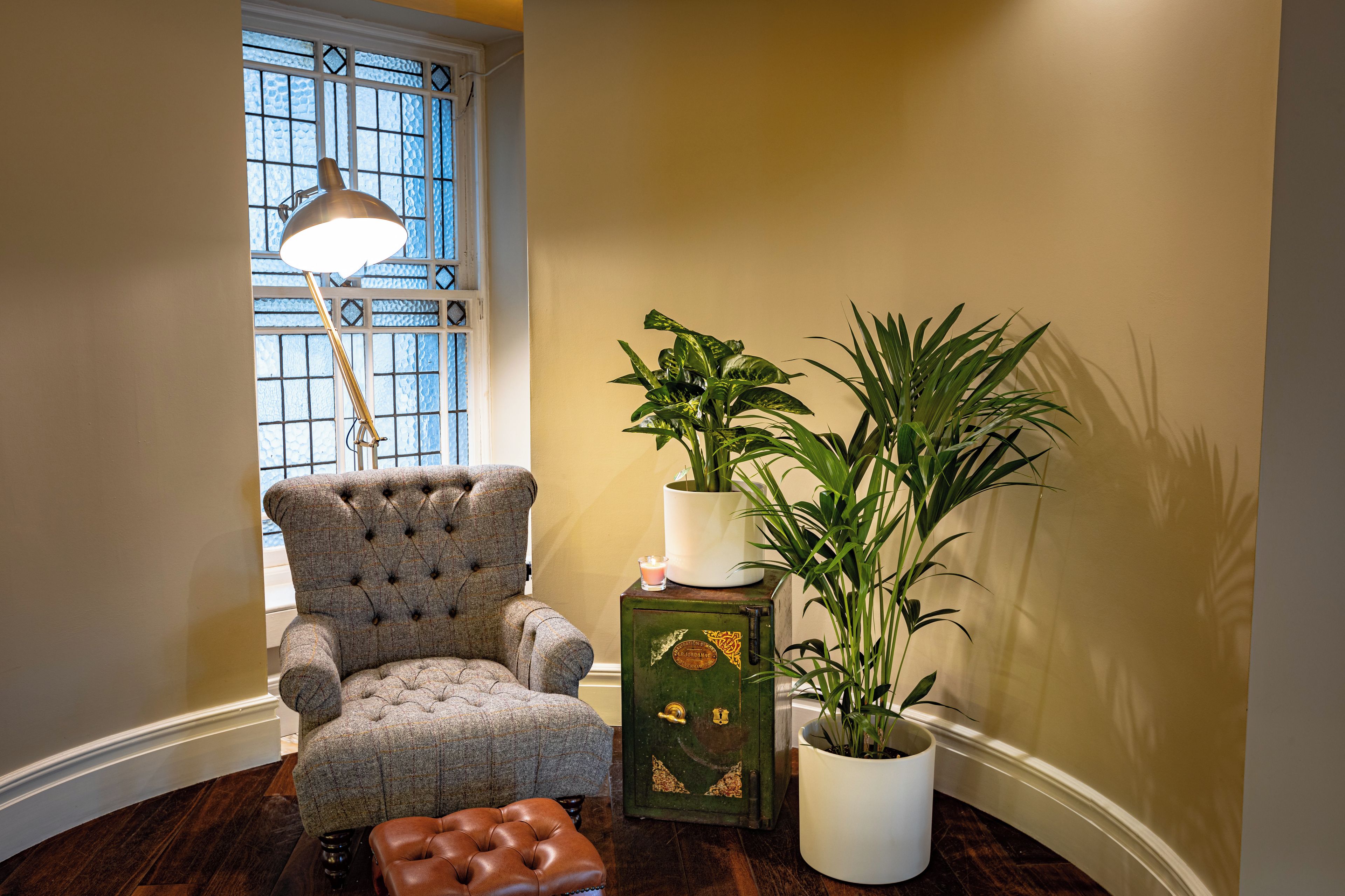 Una habitación con un sillón capitonado, una caja fuerte de estilo vintage verde, una planta alta en maceta y una lámpara de pie cerca de un ventanal con un diseño de cristal decorativo.