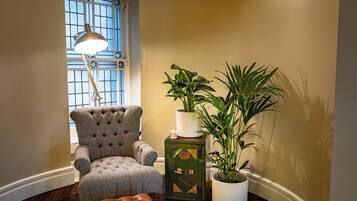 Una habitación con un sillón capitonado, una caja fuerte de estilo vintage verde, una planta alta en maceta y una lámpara de pie cerca de un ventanal con un diseño de cristal decorativo.