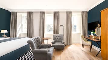 A hotel room with a bed, two armchairs, a desk with a chair, a lamp, and a TV.