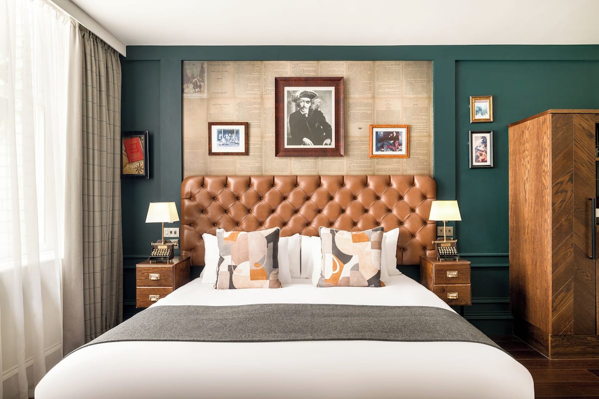 A hotel room with a tufted headboard, a large bed with patterned pillows, a wooden nightstand with a lamp, and a wardrobe.