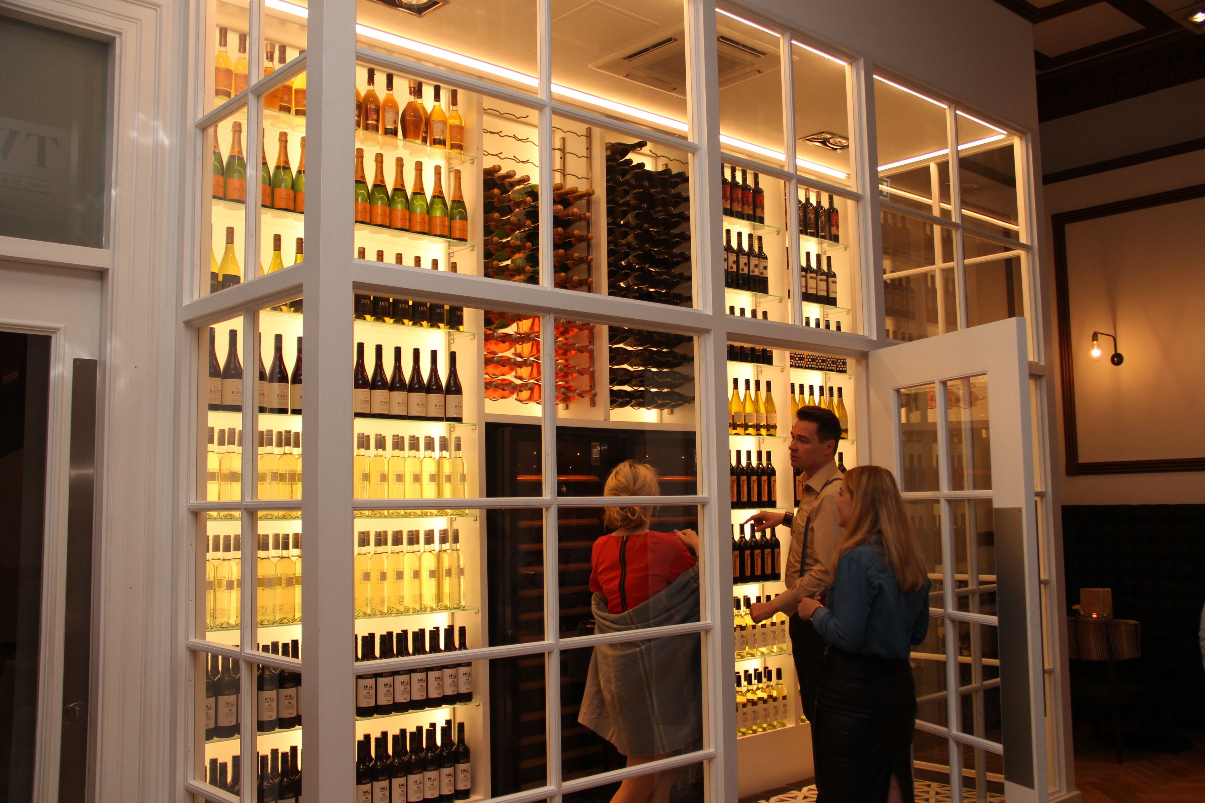 A wine cellar with shelves of bottles, a glass door entrance, and a person standing inside.