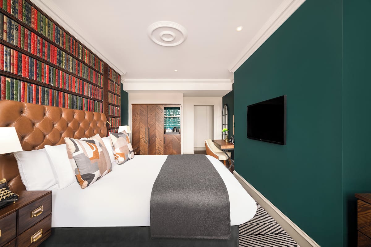 A bedroom with a large bookshelf, a tufted headboard, a flat-screen TV, and a view of a green outdoor area.