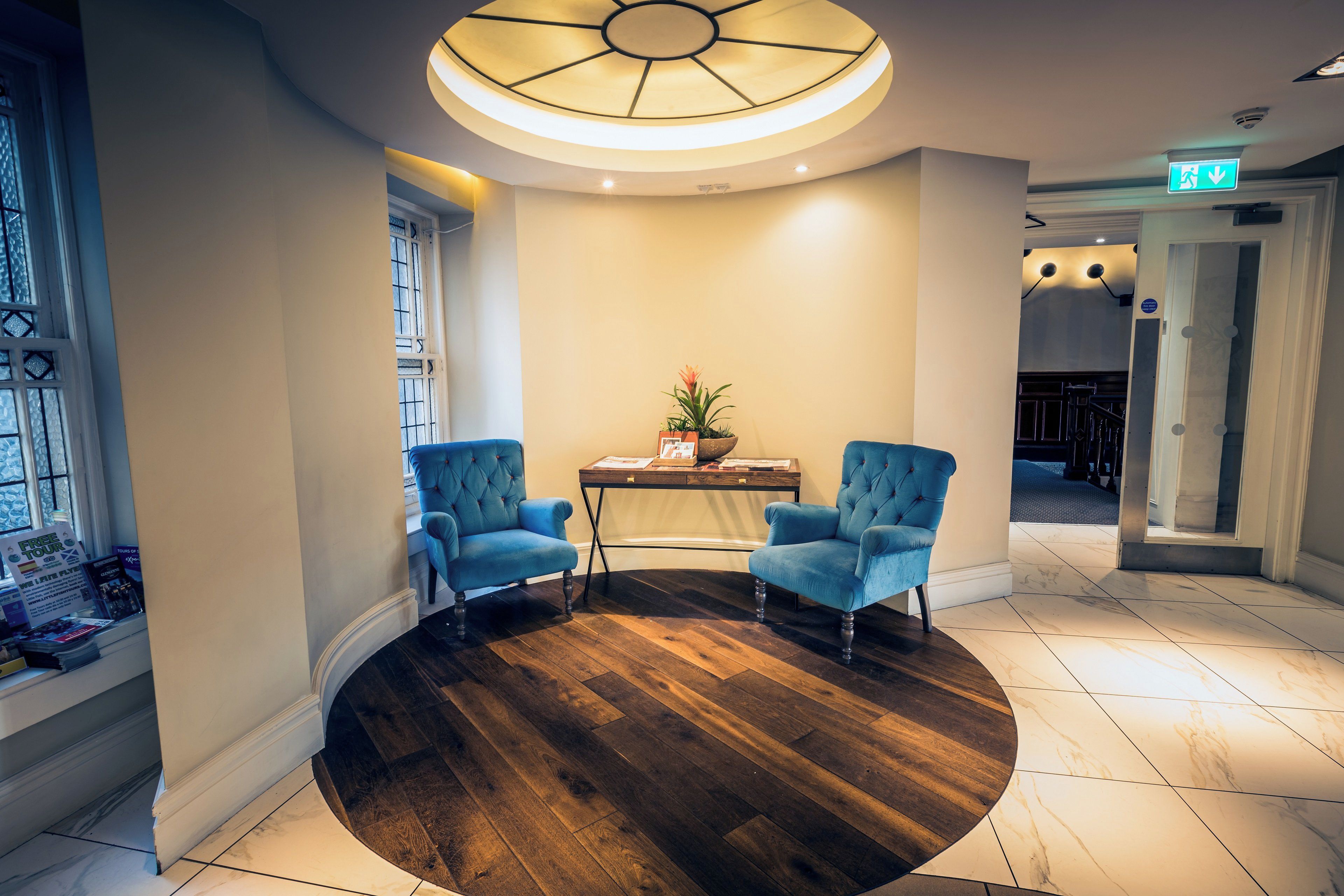 A lobby with a round wooden floor, two blue armchairs, a table with a plant, and a ceiling light fixture.