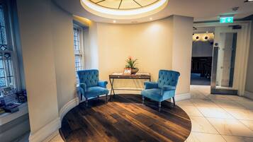 A lobby with a round wooden floor, two blue armchairs, a table with a plant, and a ceiling light fixture.
