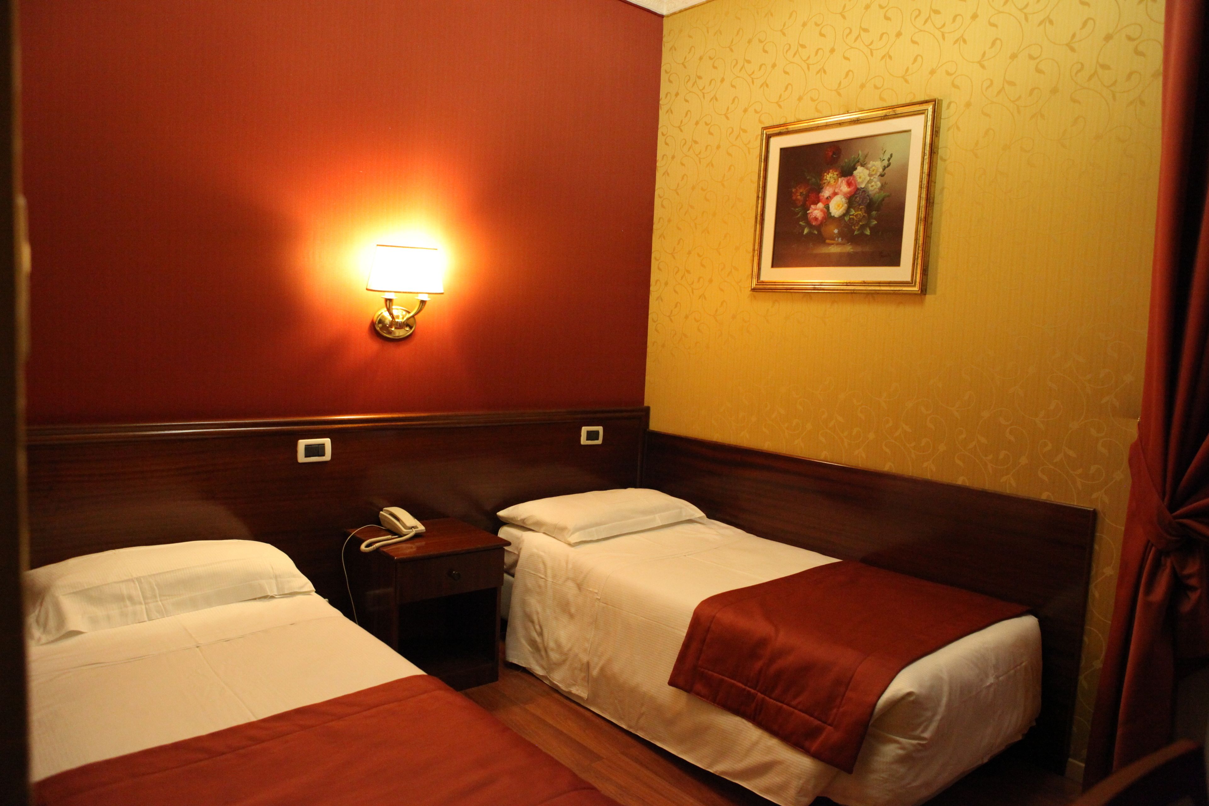Habitación de hotel con dos camas, una mesita de noche con teléfono, una lámpara de pared y un cuadro enmarco con motivos florales.