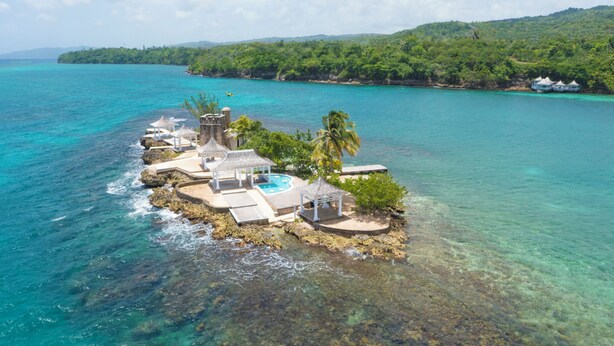 Un resort en una isla con varias cabañas sobre el agua, una piscina y abundante vegetación.