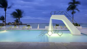 Una piscina con tobogán de agua, sillas de descanso, palmeras y vista al océano.