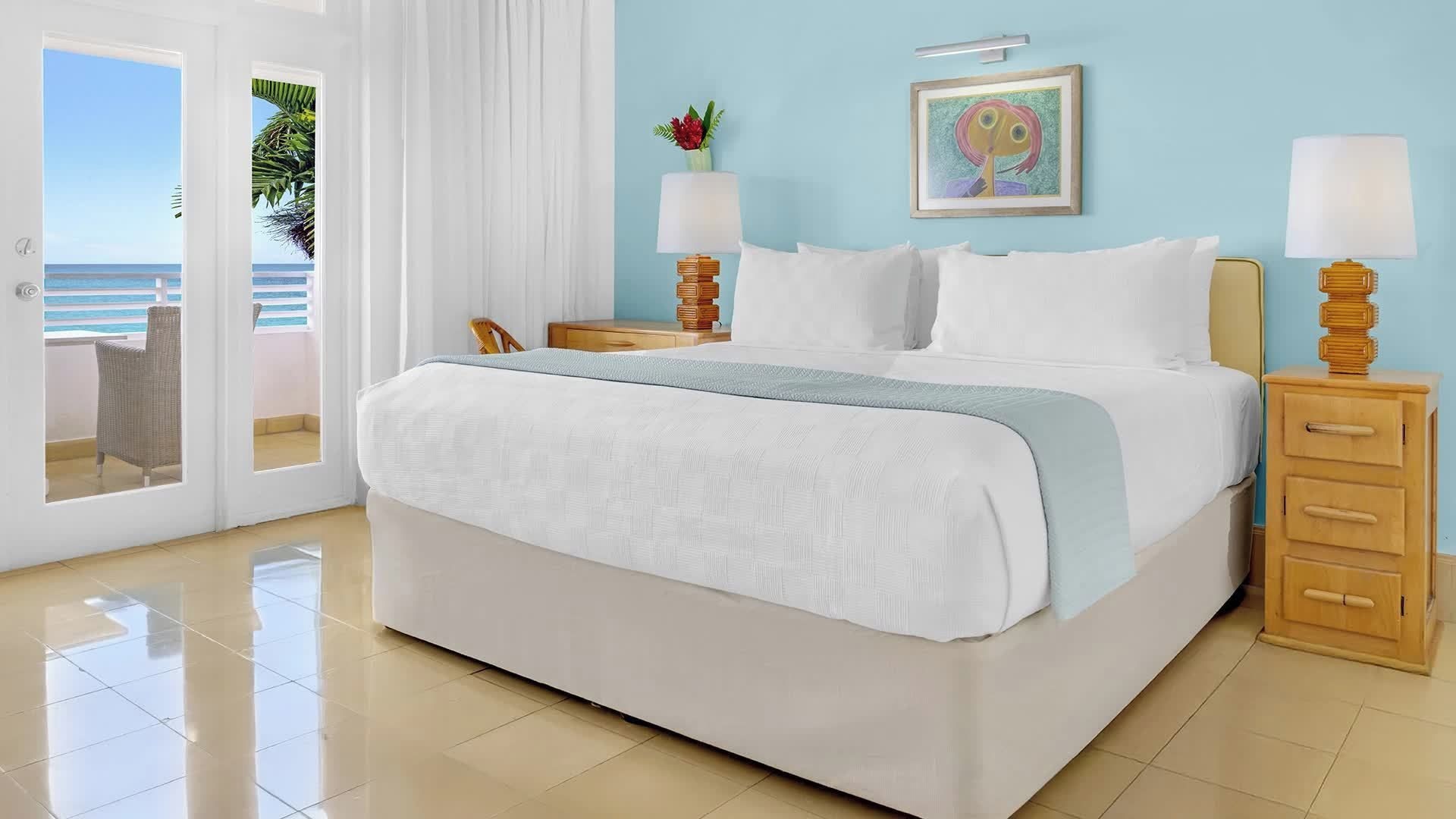 A hotel room with a bed, a nightstand, a lamp, a painting, and a view of the ocean.