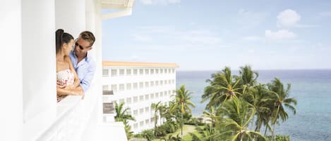 Una pareja en un balcón con vista a una piscina y al océano, con un edificio de resort al fondo.
