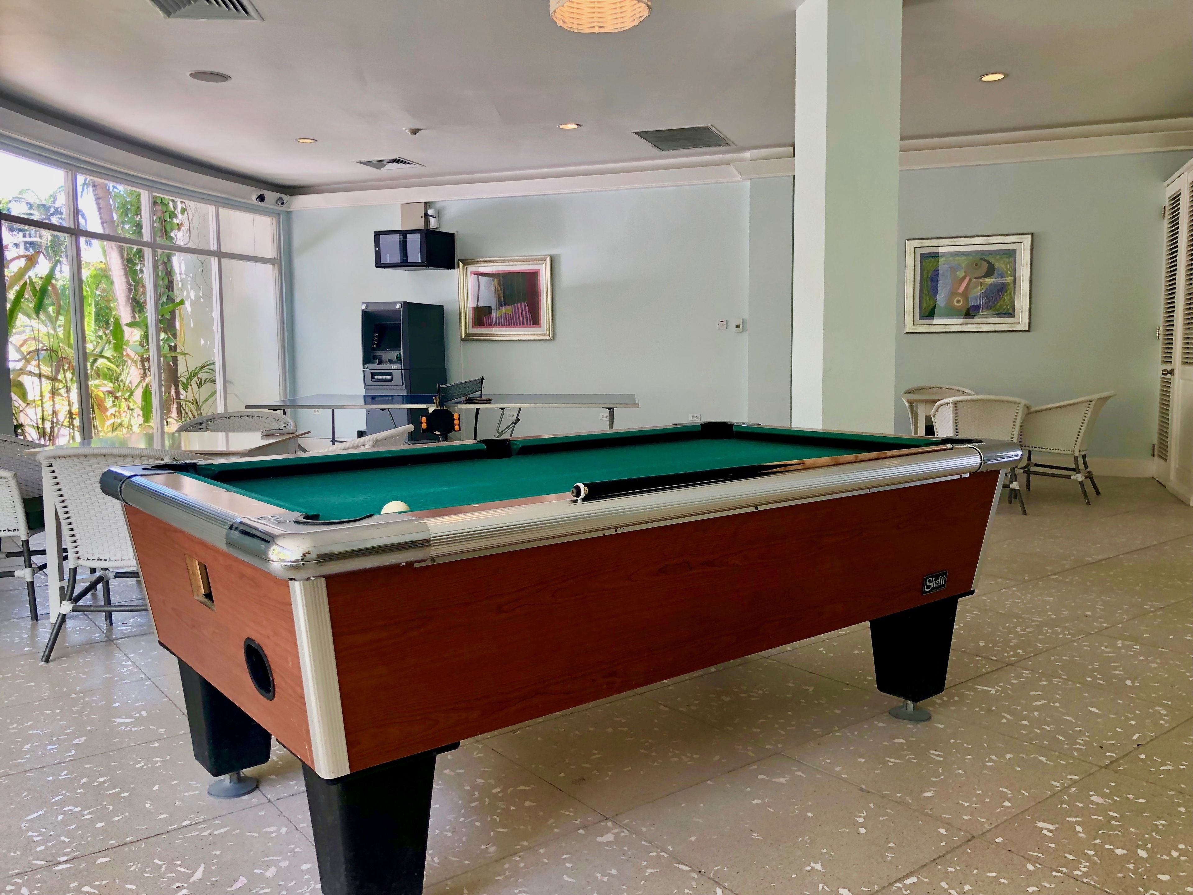 A pool table in a room with a large window, a framed picture on the wall, and a chair.