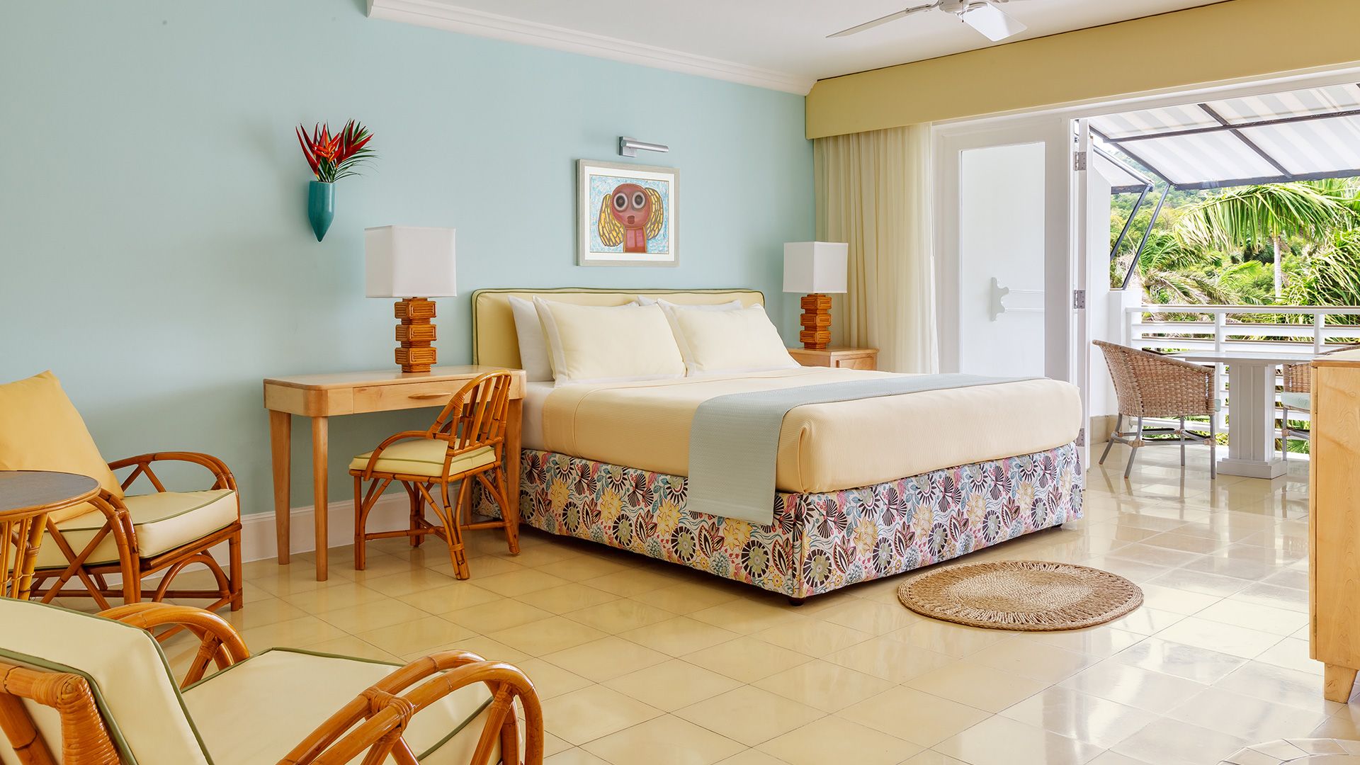 A hotel room with a bed, a desk, two chairs, a small table, a window with a view of greenery, and a ceiling fan.