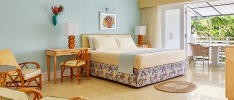 A hotel room with a bed, a desk, two chairs, a small table, a window with a view of greenery, and a ceiling fan.
