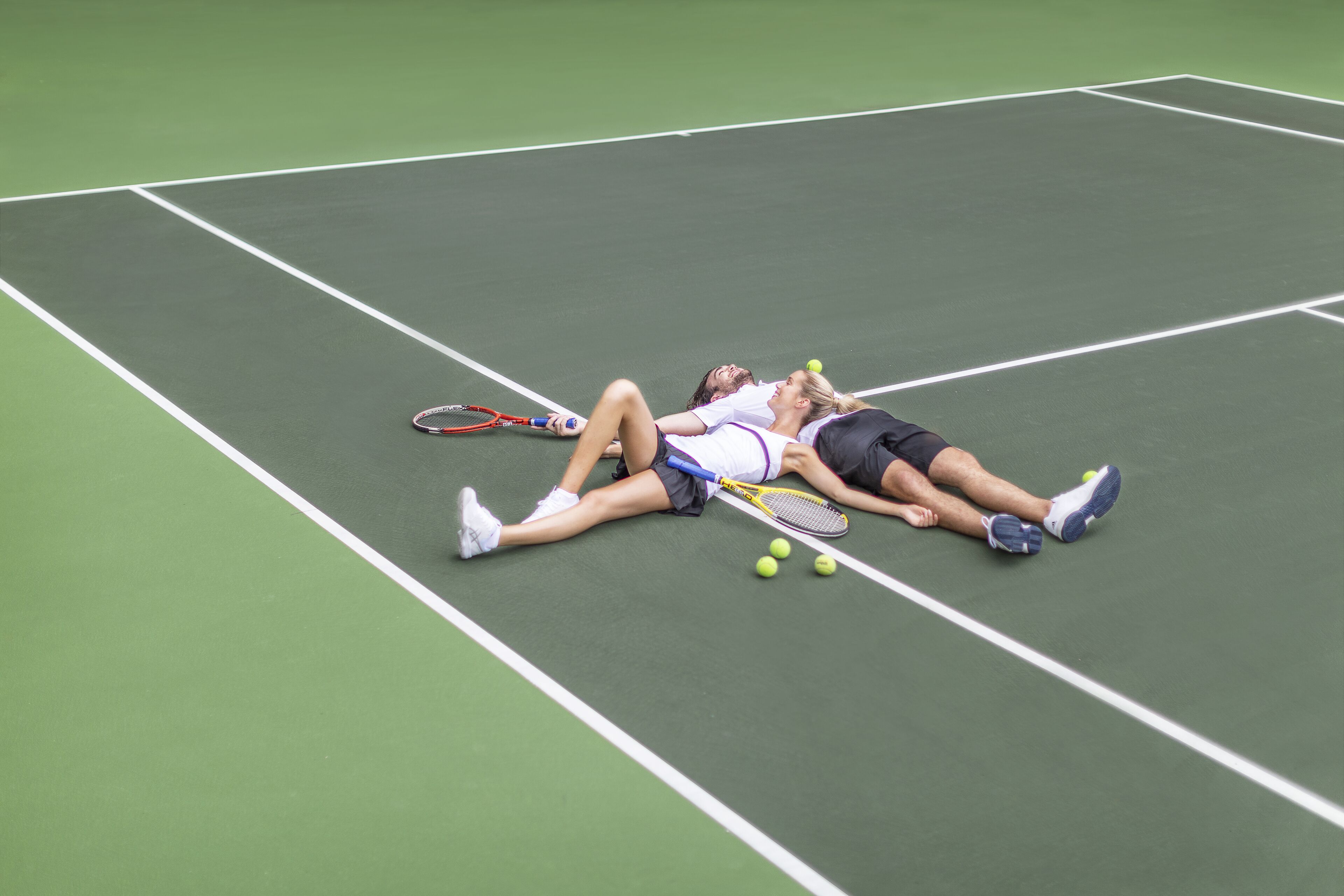 A tennis court with two players lying on the ground, tennis rackets, and tennis balls.