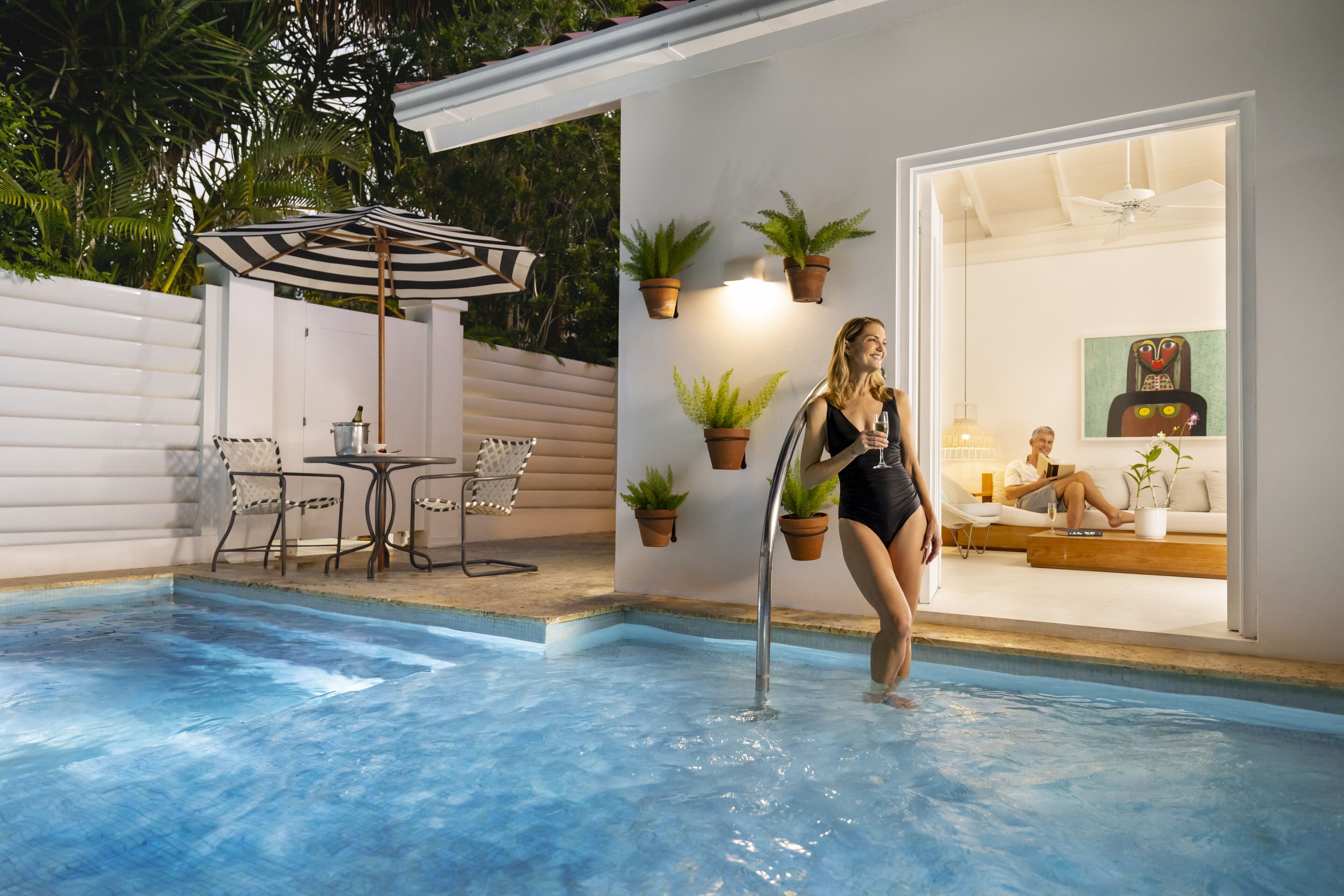A swimming pool area with a lounge chair, a table, and potted plants.