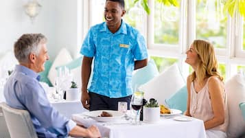 A hotel dining area with tables set for guests, a waiter in uniform, and a view of greenery outside.