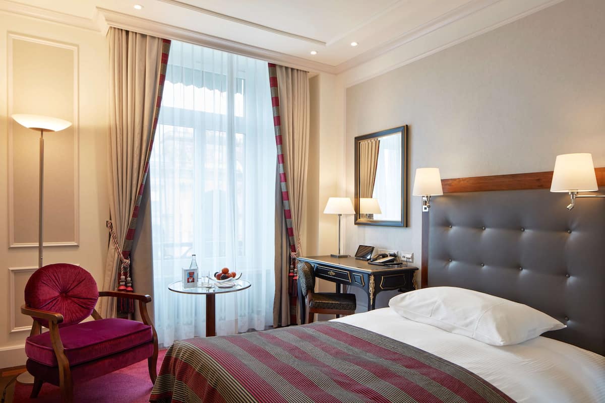 A hotel room with a bed, a desk, a chair, a small table, and a window with curtains.