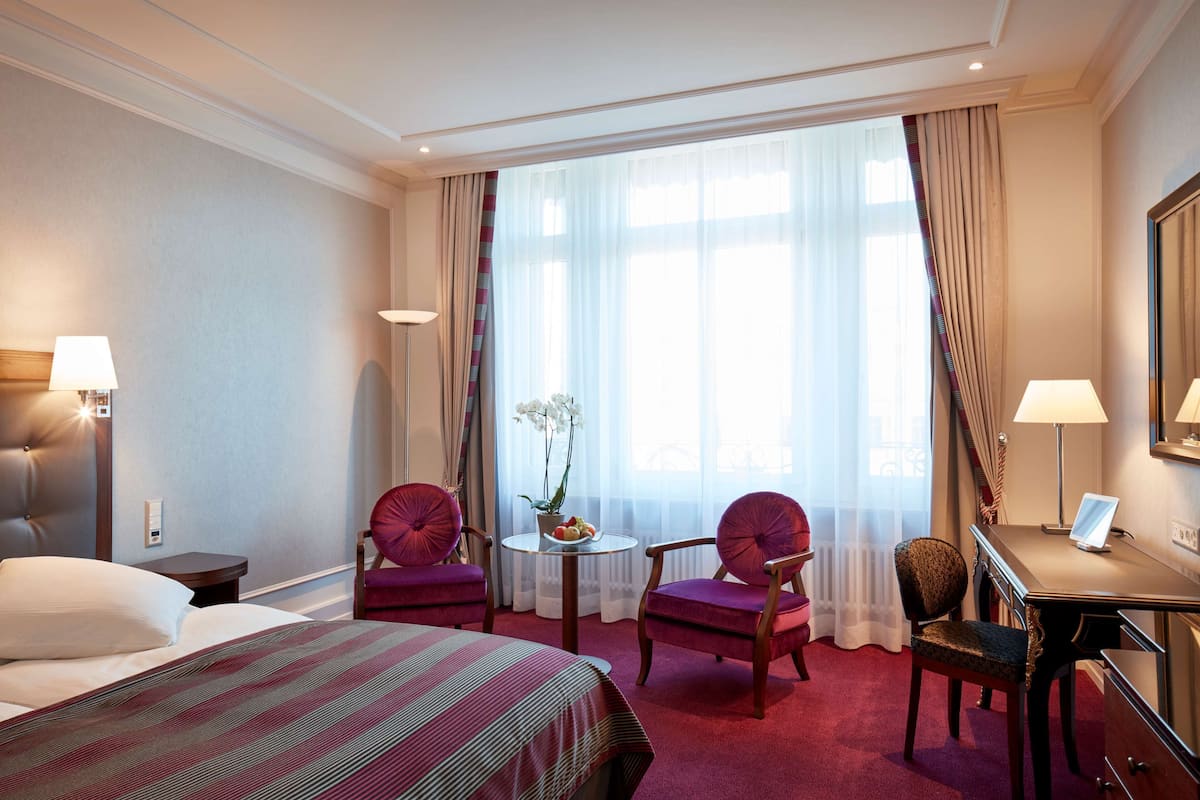 A hotel room with a bed, two chairs, a desk, a lamp, a small table with a plant, and a large window with curtains.