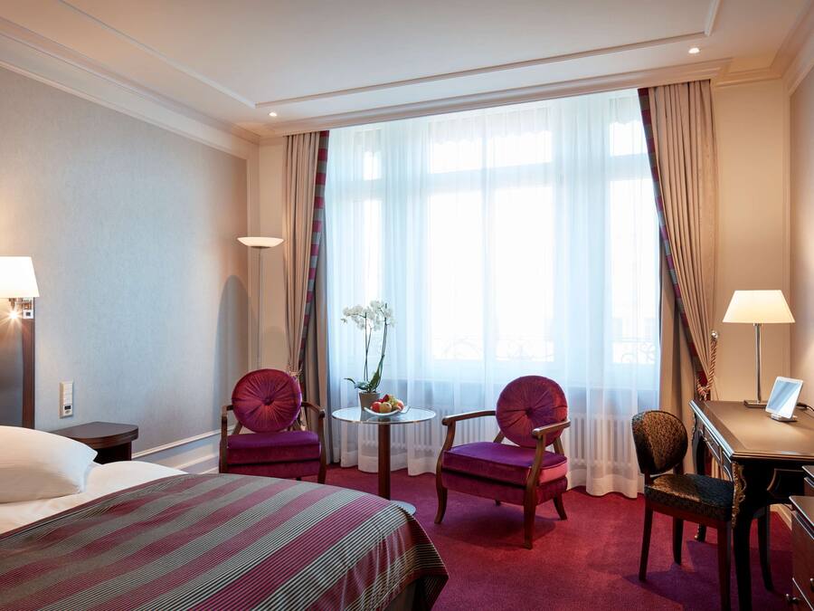 Habitación de hotel con cama, dos sillas, escritorio, lámpara, una mesita con una planta y un ventanal grande con cortinas.
