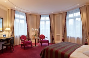 A hotel room with a large bed, two armchairs, a desk with a lamp, and a small table with flowers.