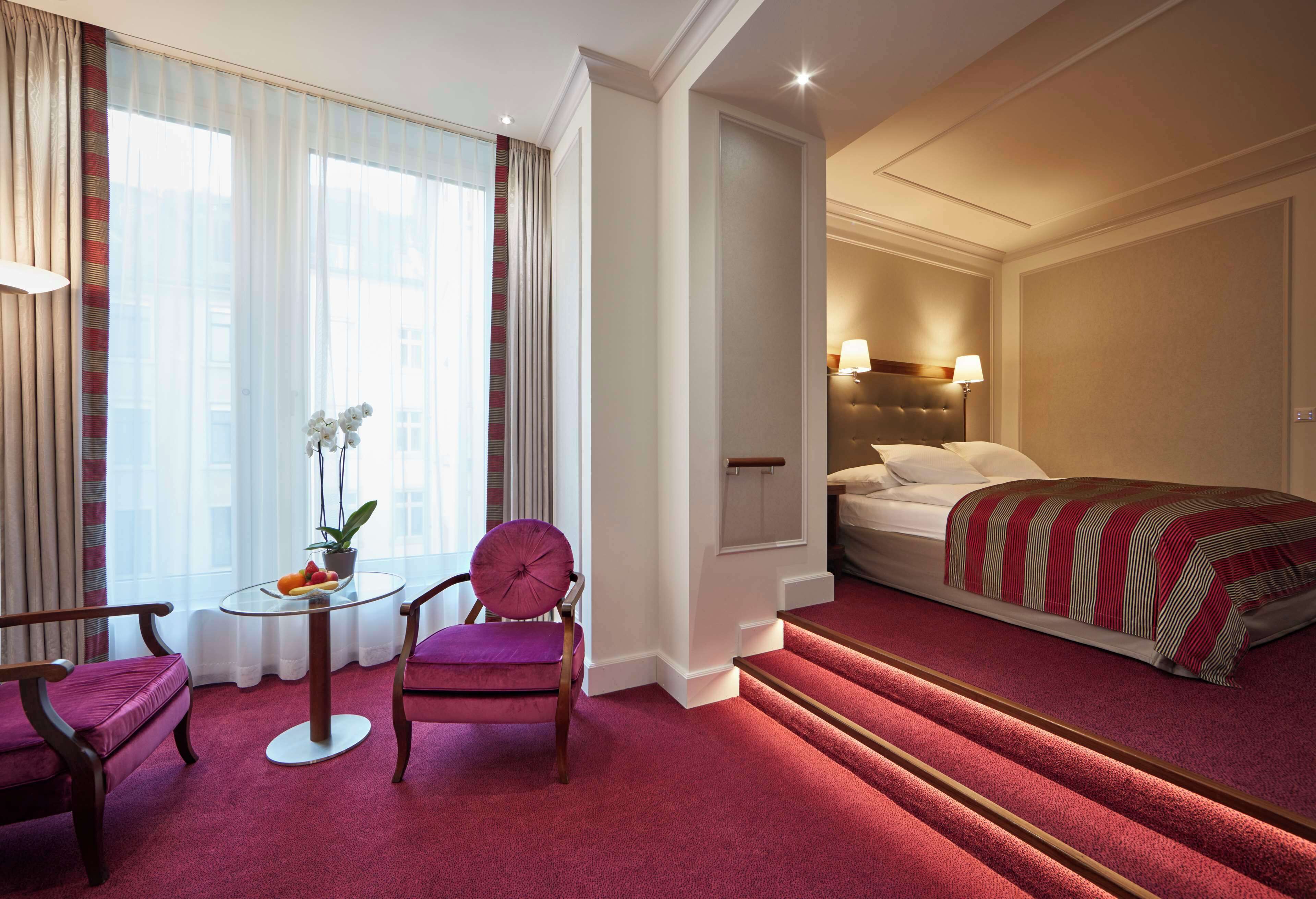 Ein Hotelzimmer mit Bett, Sessel, kleinem Tisch mit Pflanze und Fenster mit Vorhängen.