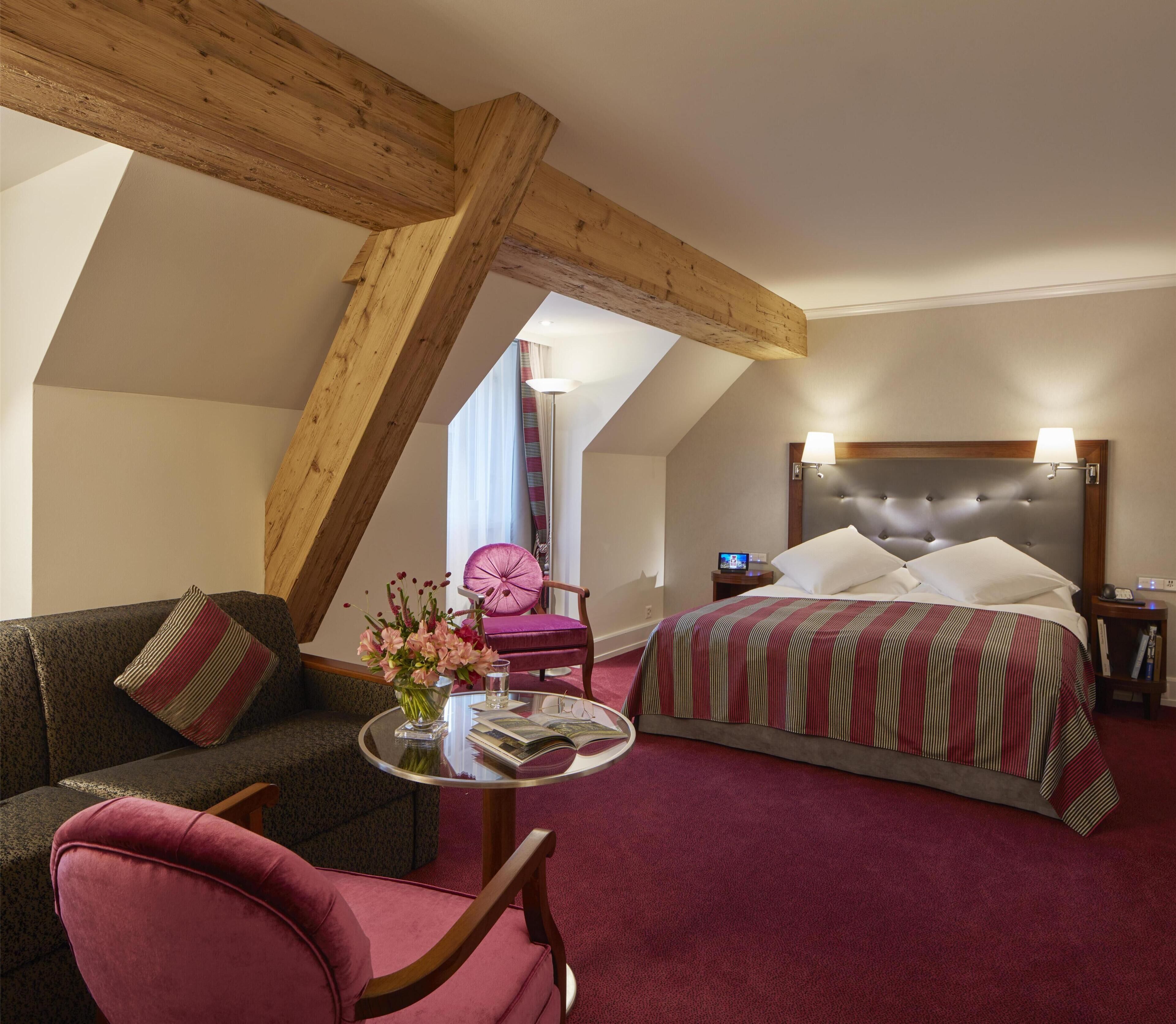 A hotel room with a bed, a sofa, a chair, a small table with a vase of flowers, and a window with curtains.