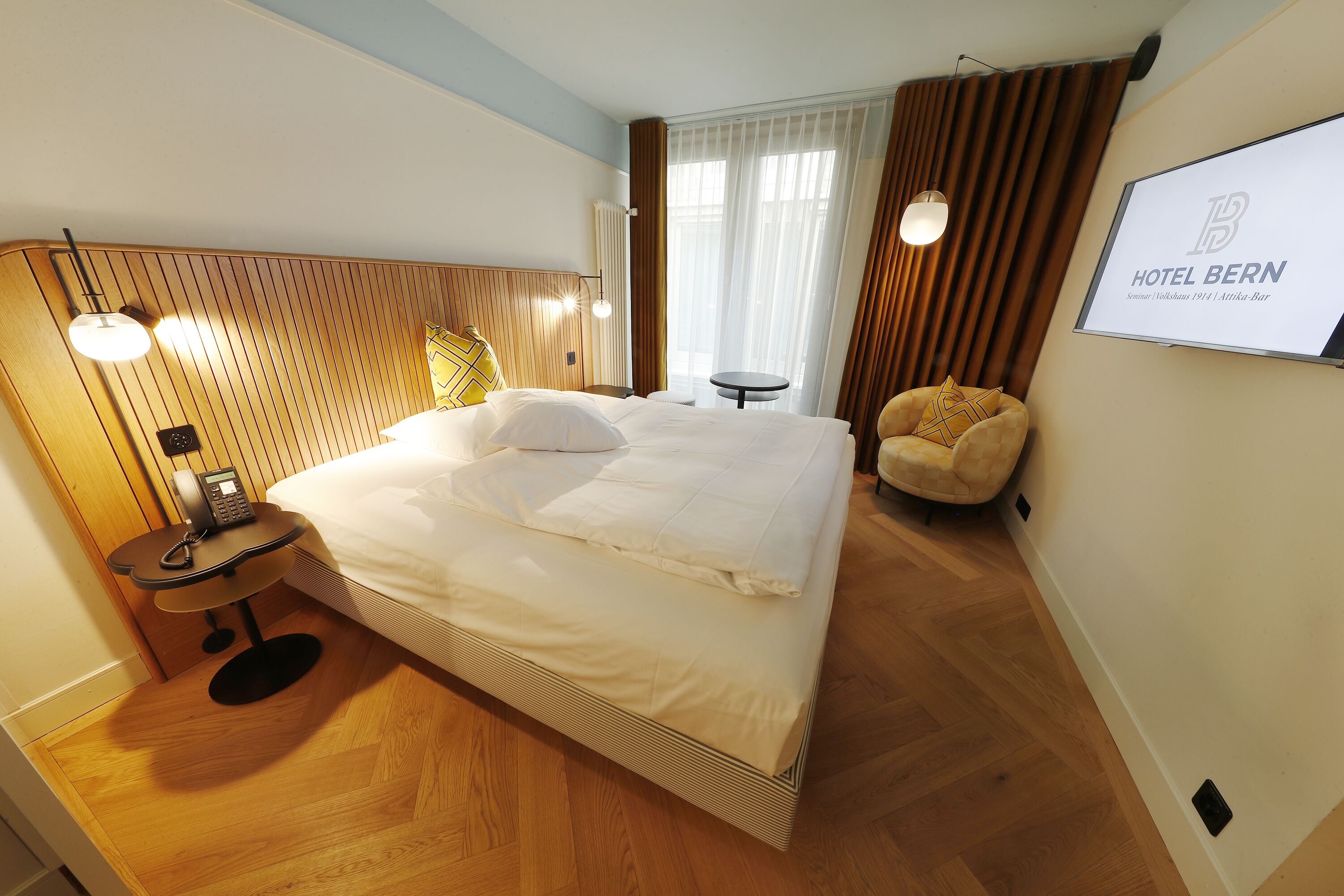 A hotel room with a bed, bedside table with a phone, a small table, a chair, and a TV on the wall.