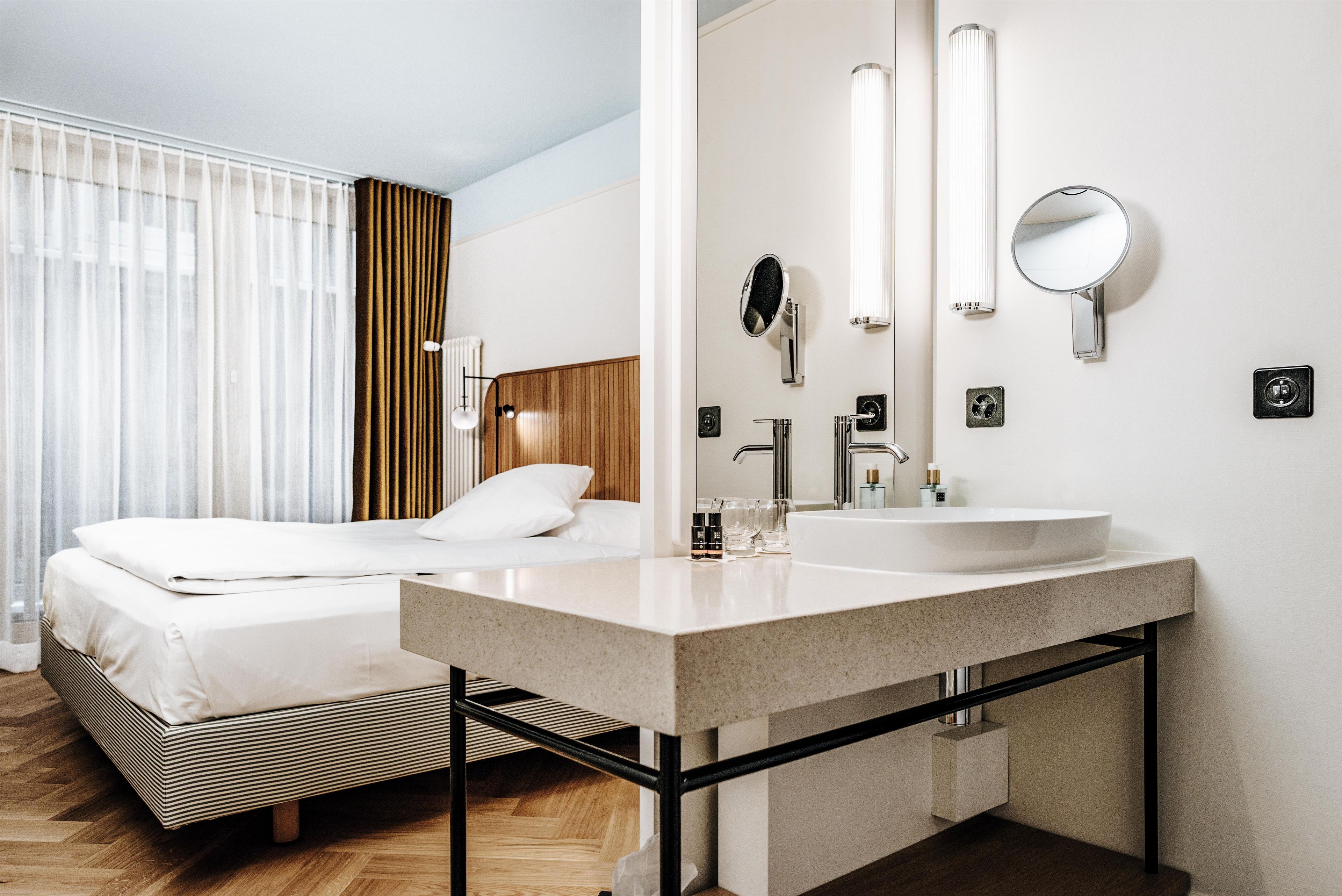 Habitación de hotel con cama, un espejo grande, lavamanos doble y ventana con cortinas.