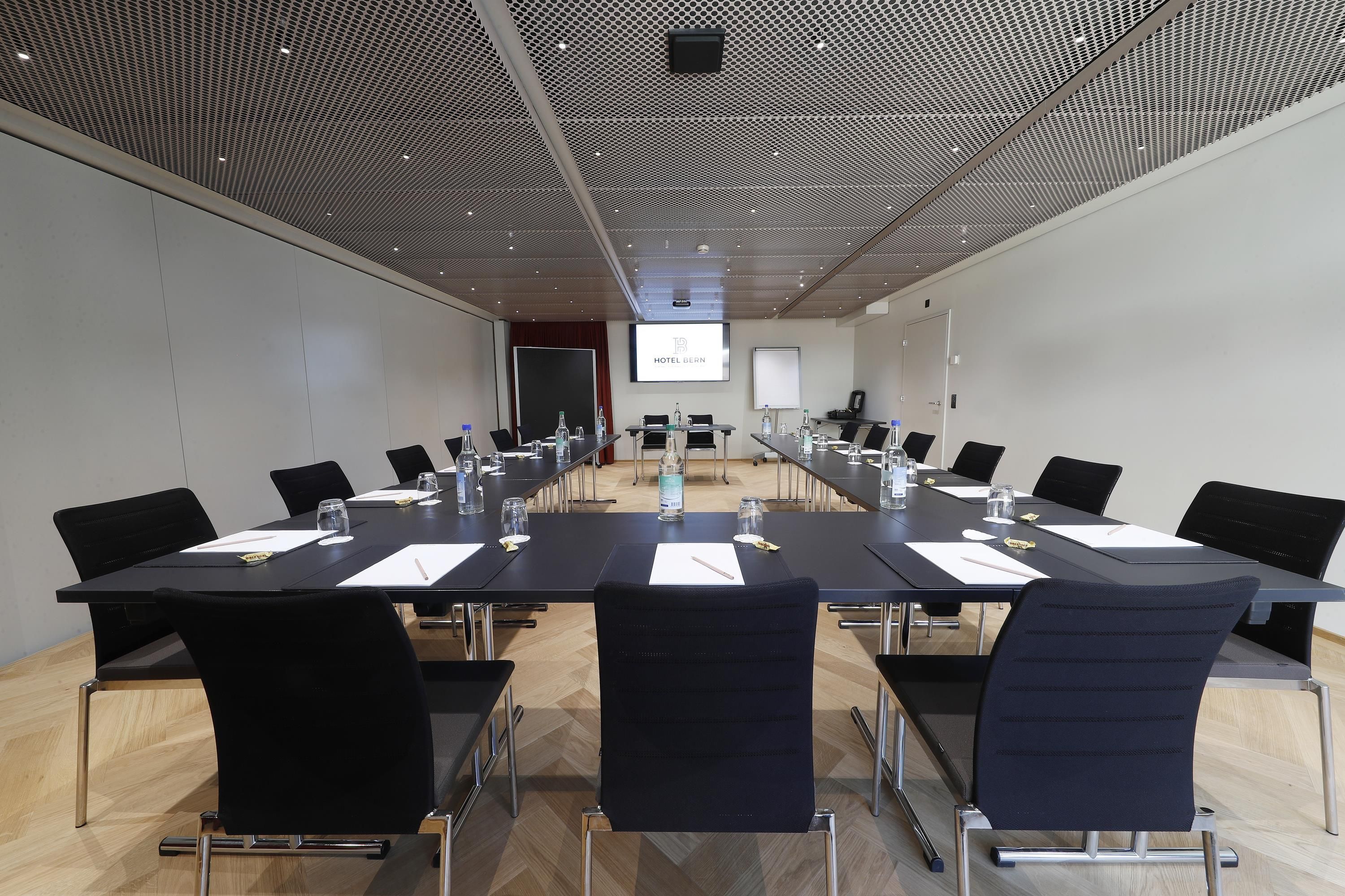 Ein Konferenzraum mit einem langen Tisch, schwarzen Stühlen und einer Projektionsleinwand, auf der 'HOTEL GEN' angezeigt wird.