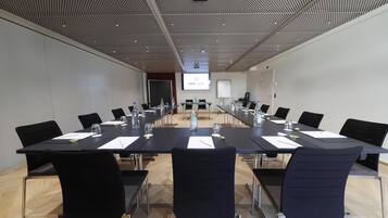 Ein Konferenzraum mit einem langen Tisch, schwarzen Stühlen und einer Projektionsleinwand, auf der 'HOTEL GEN' angezeigt wird.
