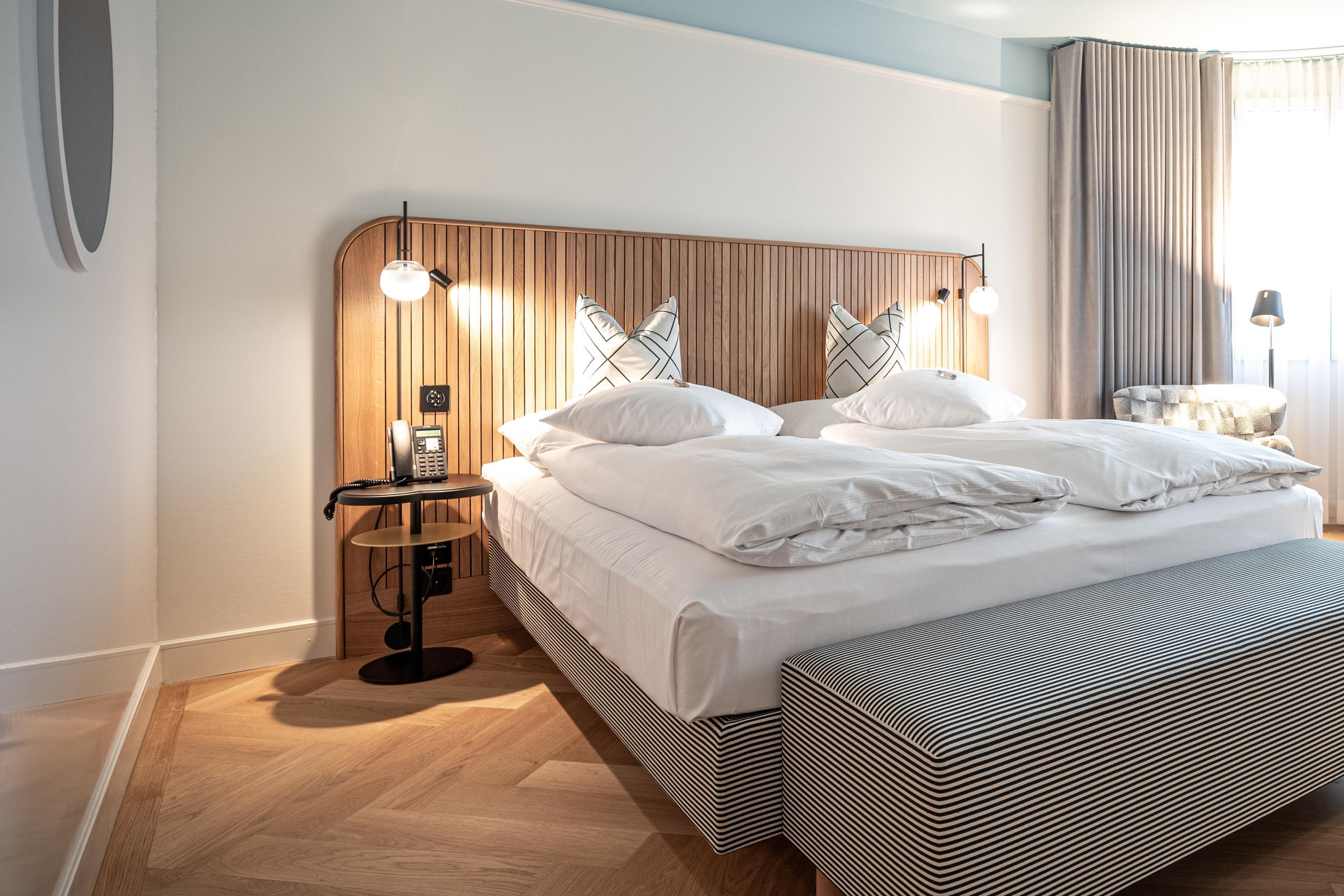 Ein modernes Hotelzimmer mit einem Holzkopfteil, einem großen Bett mit weißer Bettwäsche, einem Nachttisch mit Lampe und einem kleinen runden Tisch mit Telefon.