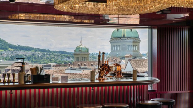 A bar with stools, a view of a cityscape, and a chandelier.