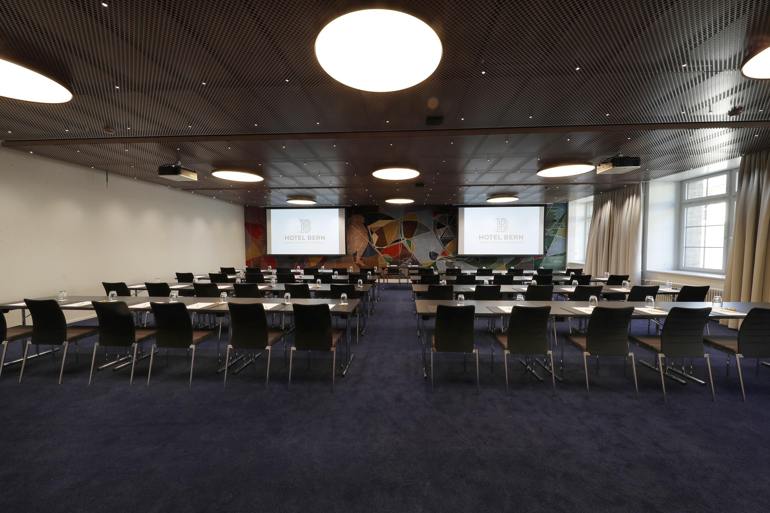 Ein Konferenzraum mit mehreren Tischen und Stühlen, großen Bildschirmen, die den Hotelnamen zeigen, und einem modernen Deckengestaltungsstil.