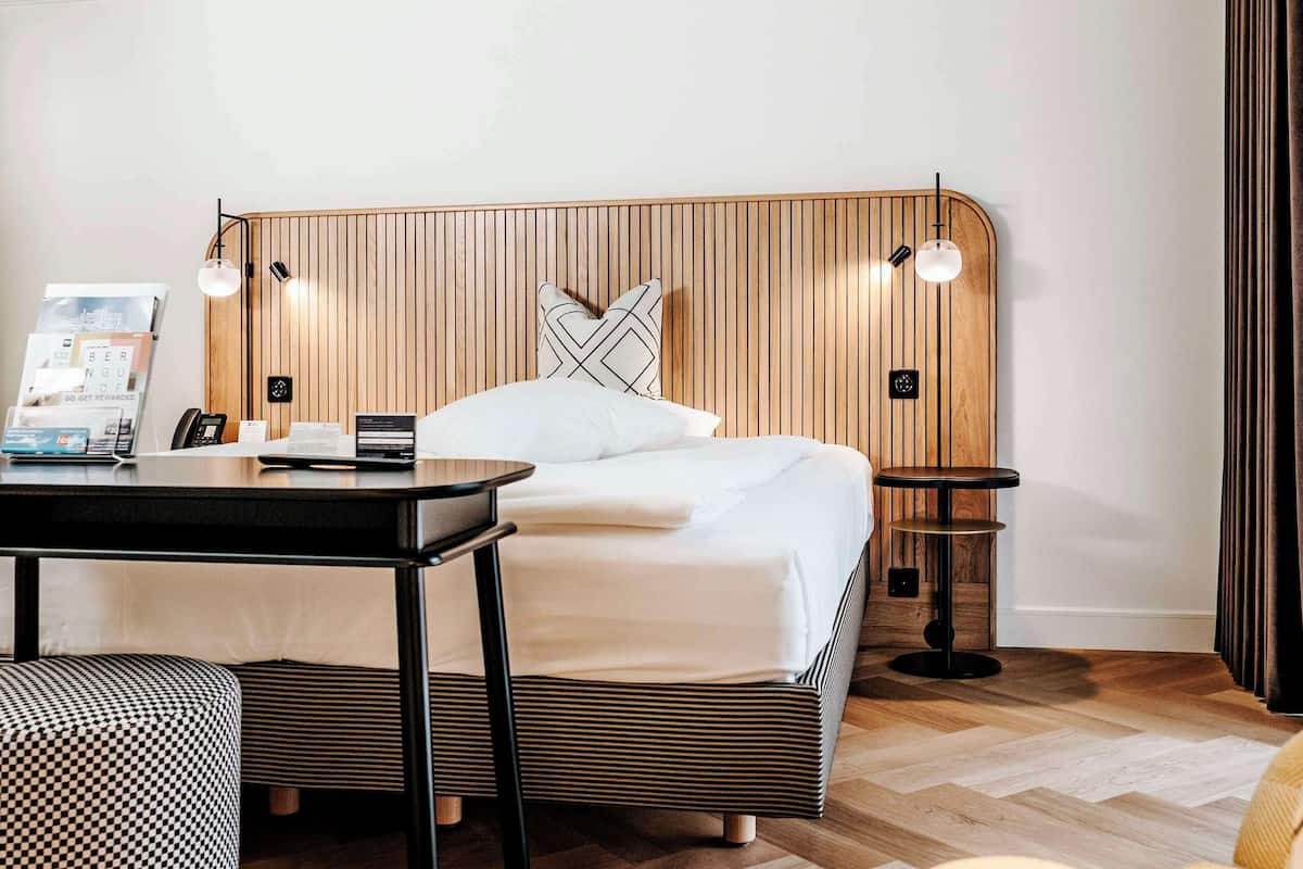 A modern hotel room with a wooden headboard, a bed, a desk with a lamp, a round table, and a patterned ottoman.