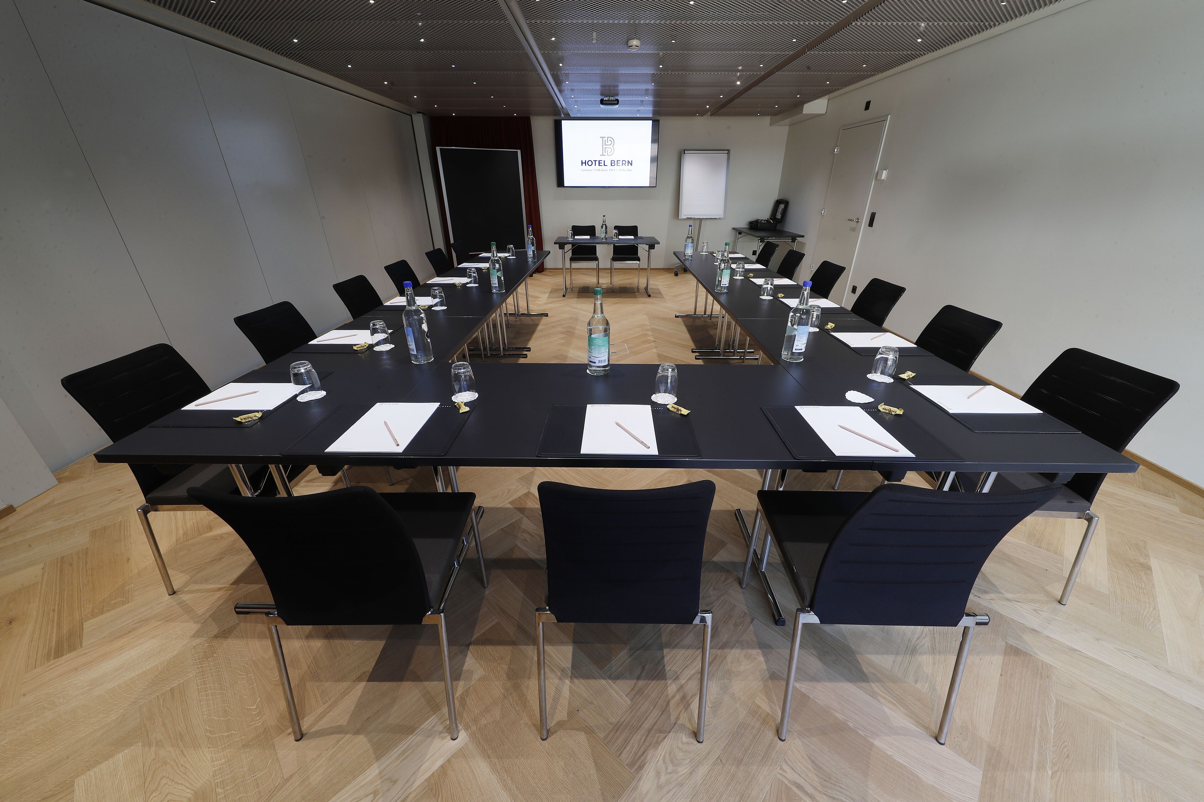 A conference room with a long table, black chairs, water bottles, and a projector screen displaying "HOTEL BERN".