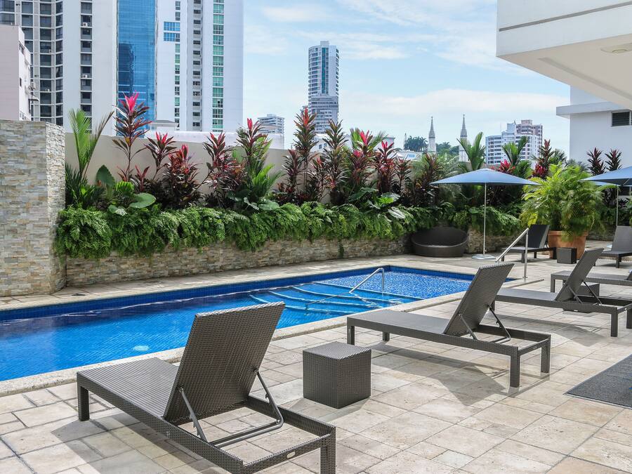 Zona de piscina en la azotea con sillones de descanso y sombrillas, rodeada de exuberante vegetación y edificios altos al fondo.