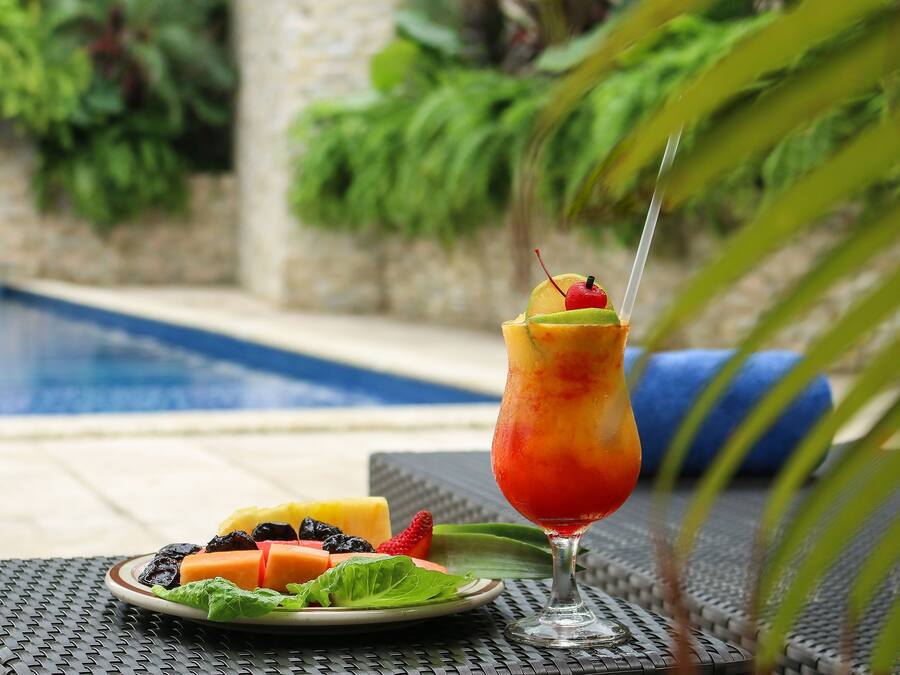 Un décor tropical avec une piscine, une assiette de fruits et un verre à cocktail.