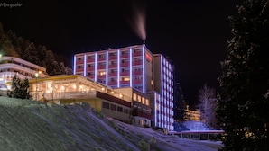 A multi-story hotel with illuminated windows and balconies, surrounded by snow-covered terrain and trees.