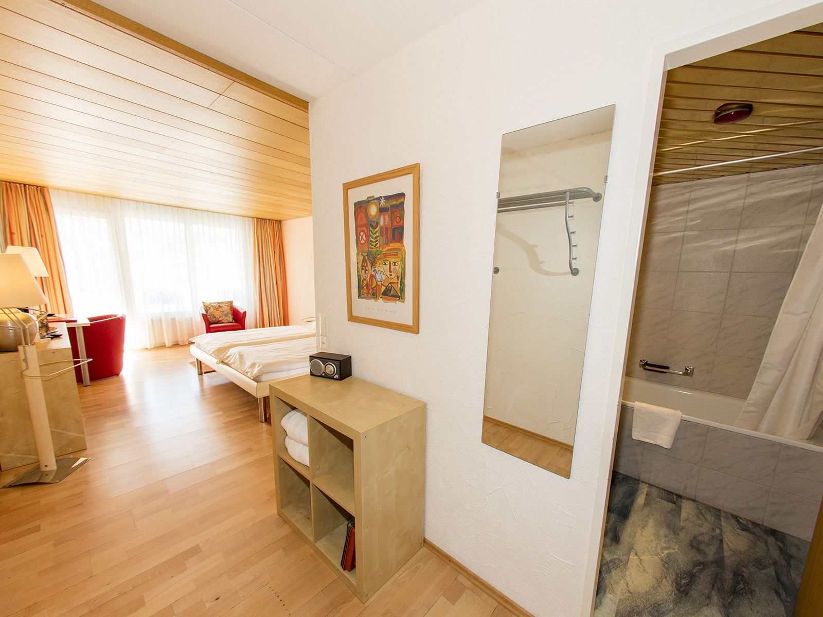 A hotel room with a bed, a bedside table with a radio, a mirror, a shower area, and a window with curtains.