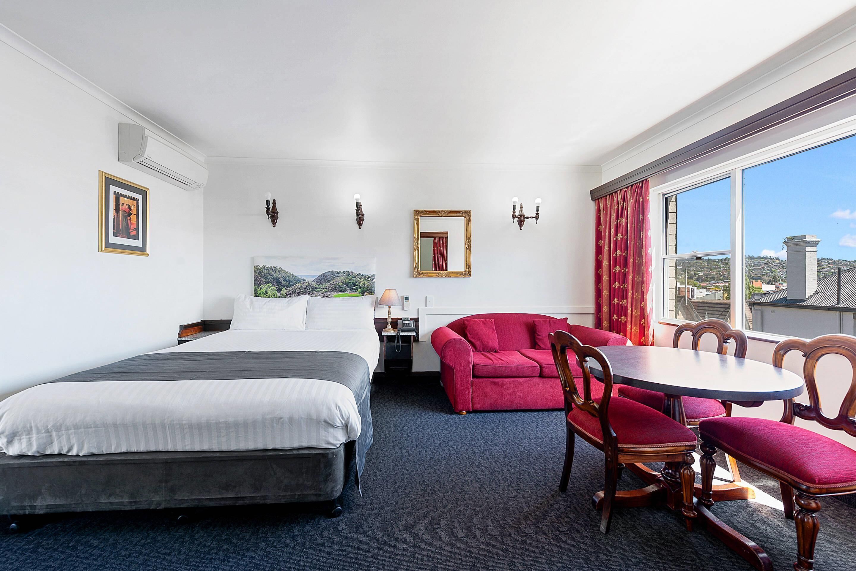 Habitación de hotel con cama, escritorio con lámpara, sofá rojo, mesa de comedor con sillas y un ventanal con vistas.