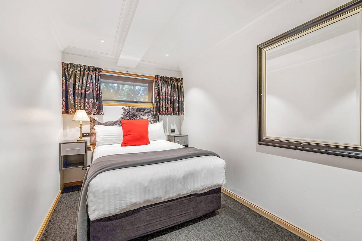 A hotel room with a bed, nightstands, a lamp, a window with curtains, and a large mirror.