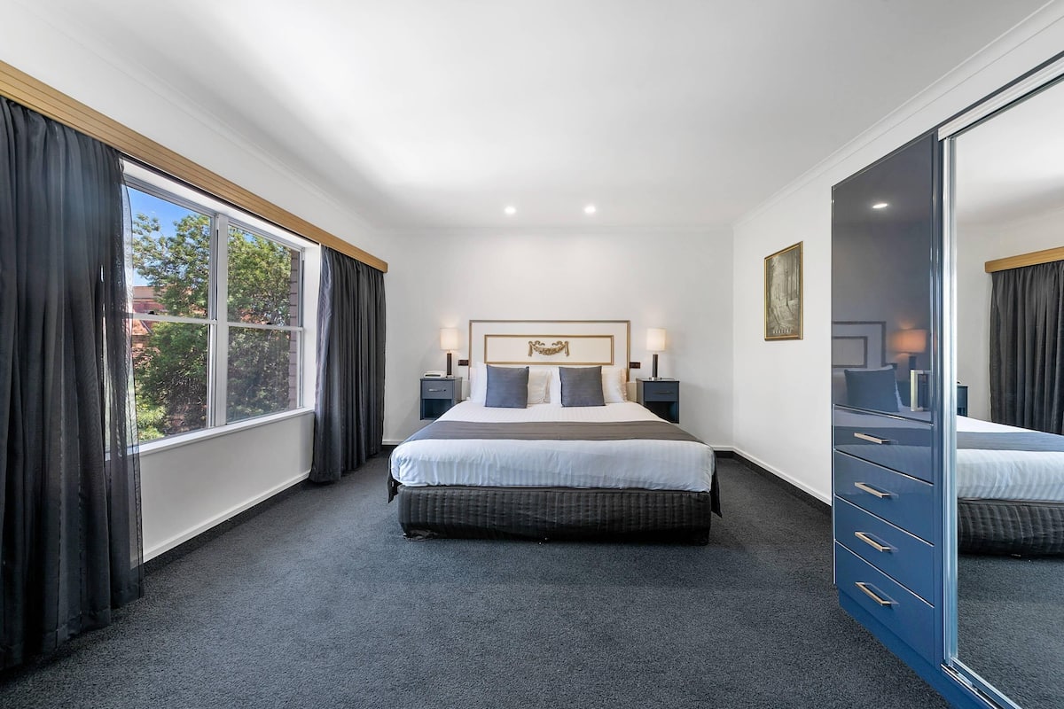 A hotel room with a large bed, a dresser, a window with curtains, and a framed picture on the wall.